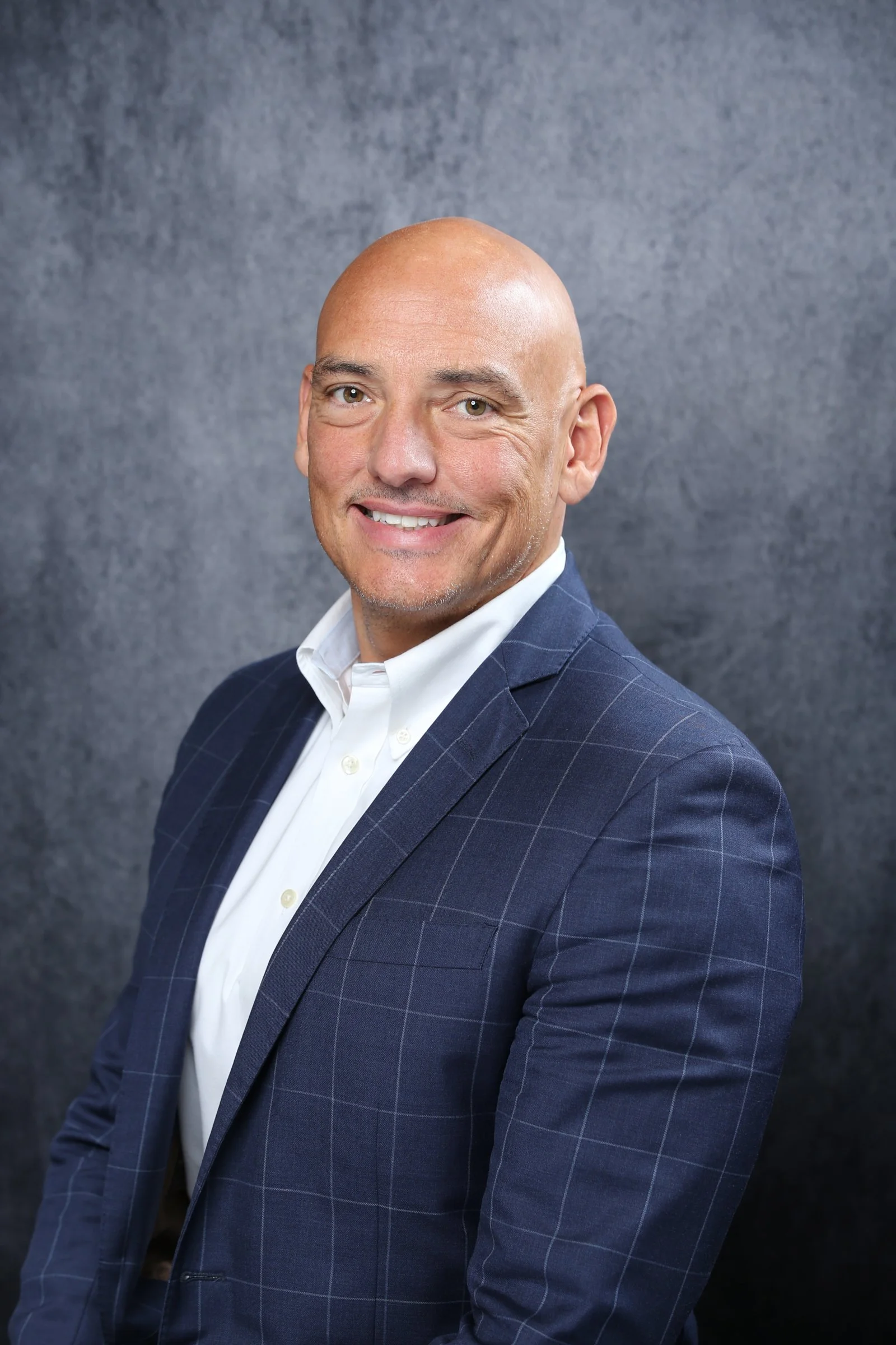 A bald man wearing a navy blue checkered suit and a white dress shirt, smiling, against a gray textured background.