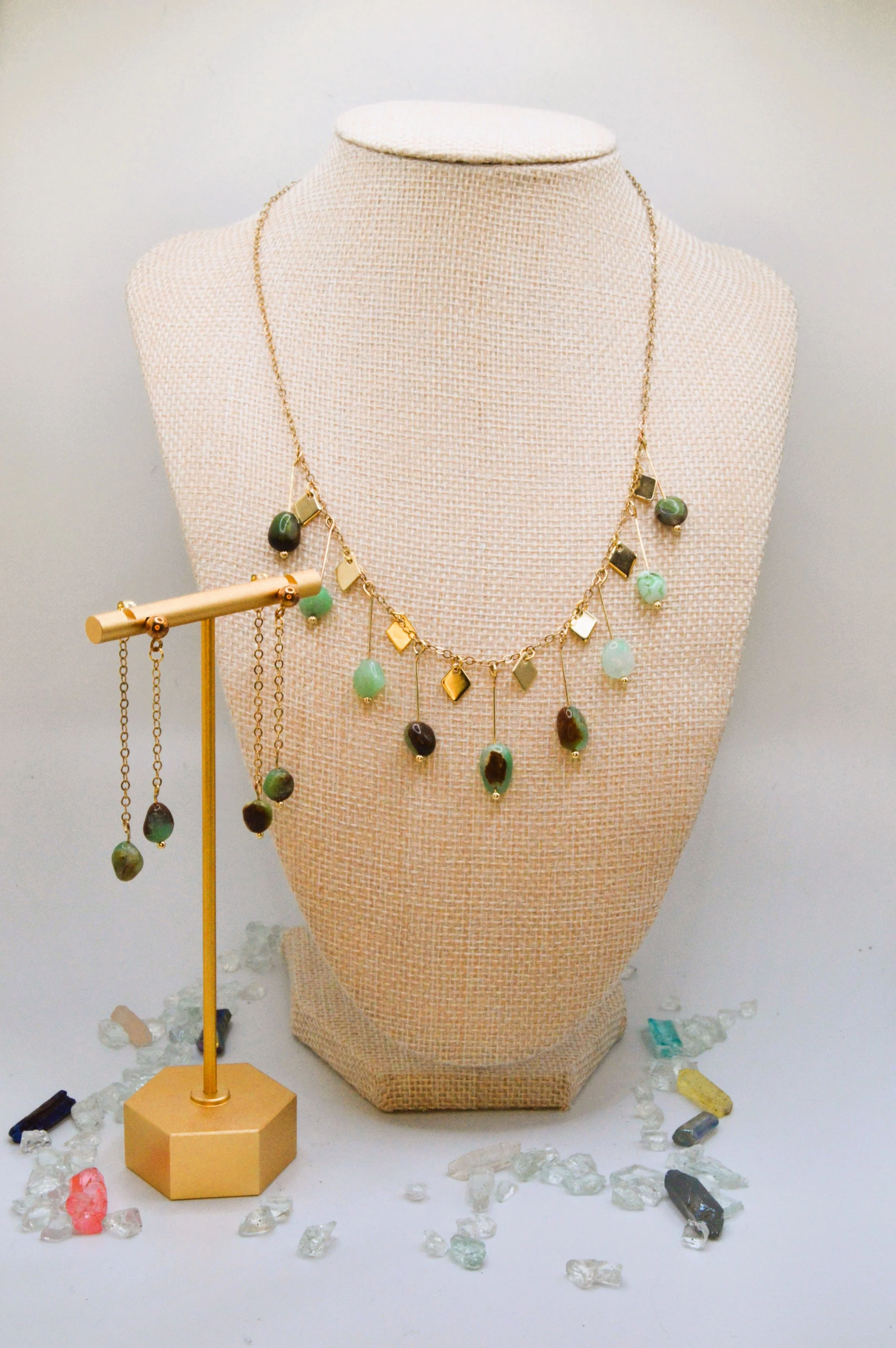 Display of jewelry including a necklace with green and black stones, earrings, and various loose gemstones and beads on a white surface with a beige jewelry bust and a gold stand.