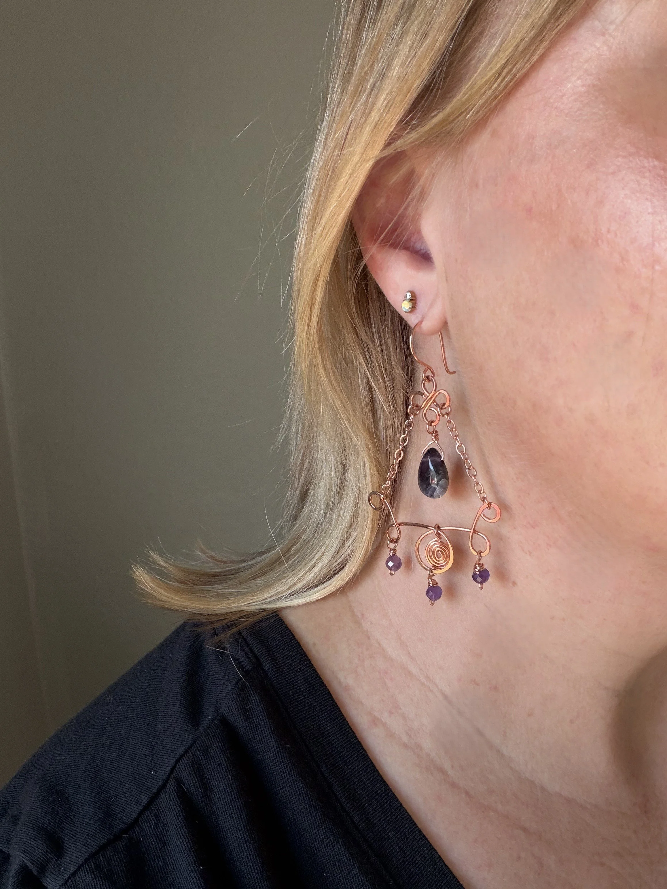 WOMAN WEARING Handmade copper chandelier earring worn on an ear, with scroll connectors, copper chains, a center spiral, an amethyst teardrop, and three small faceted amethyst beads.