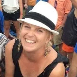 A woman smiling, wearing a white fedora hat and black top, in a crowd of people outdoors.
