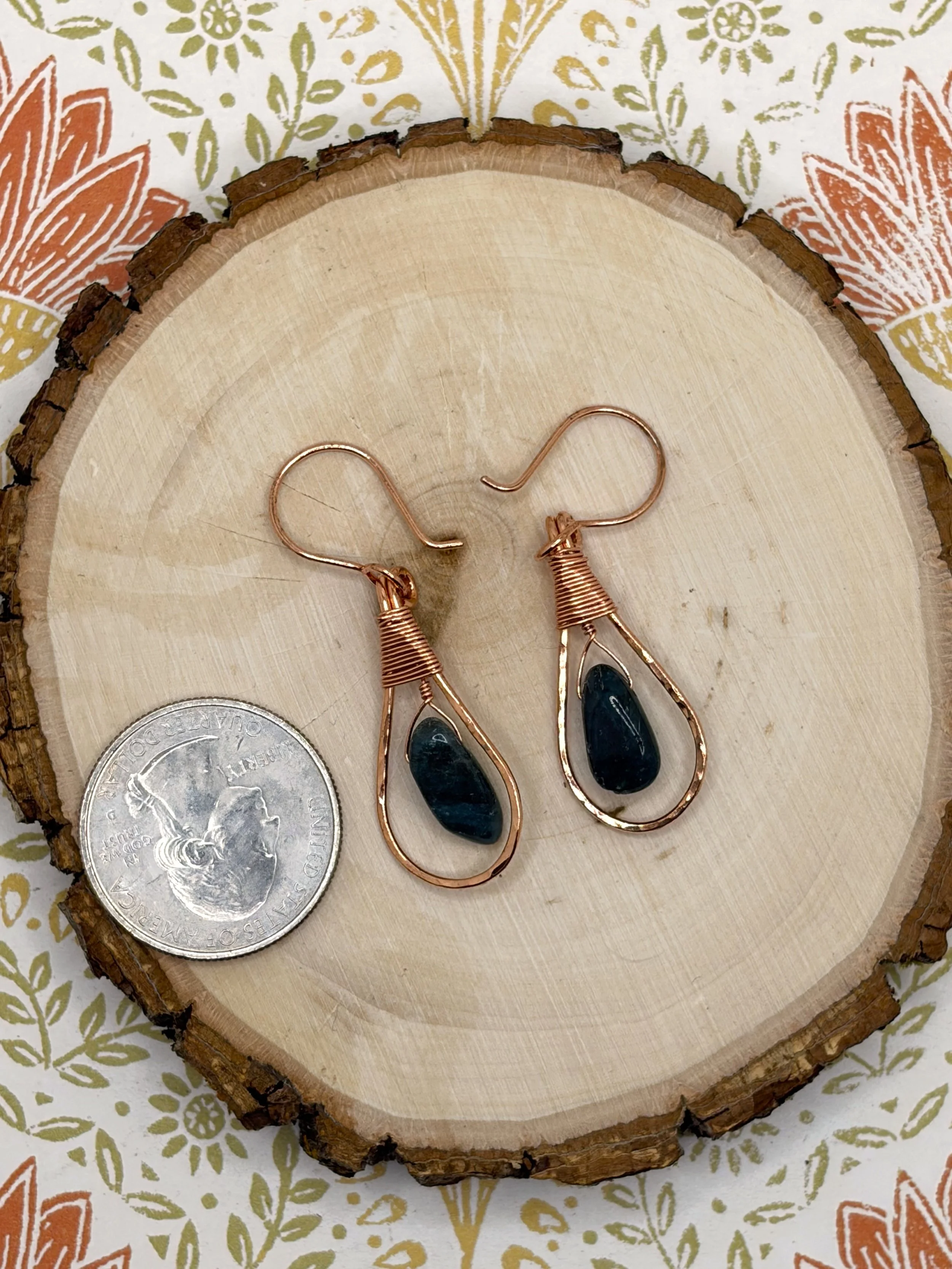 Pair of handmade hammered copper teardrop earrings with coiled wire caps and deep blue tourmaline beads on a wooden slice, shown next to a US quarter for scale.