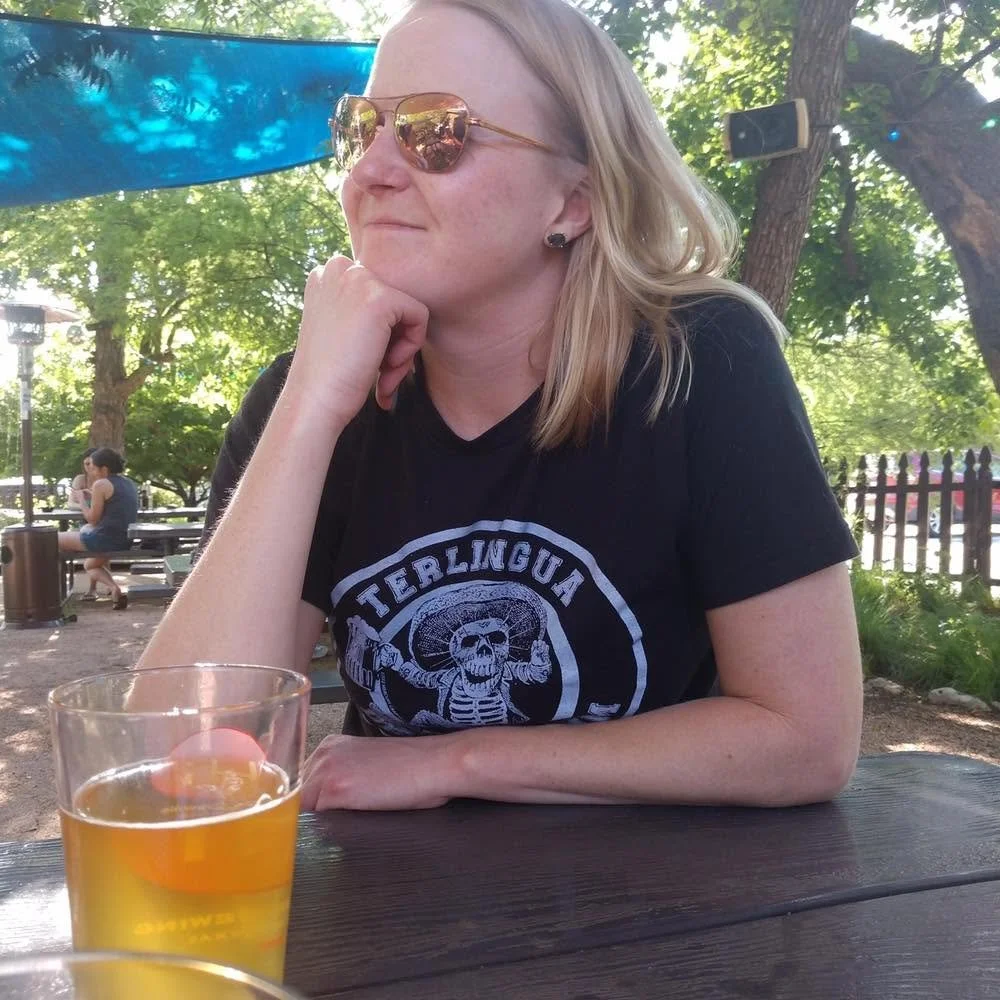 A woman with blonde hair wearing sunglasses and a black T-shirt sitting at an outdoor table with a glass of beer, resting her chin on her hand, in a park setting with trees and other people in the background.