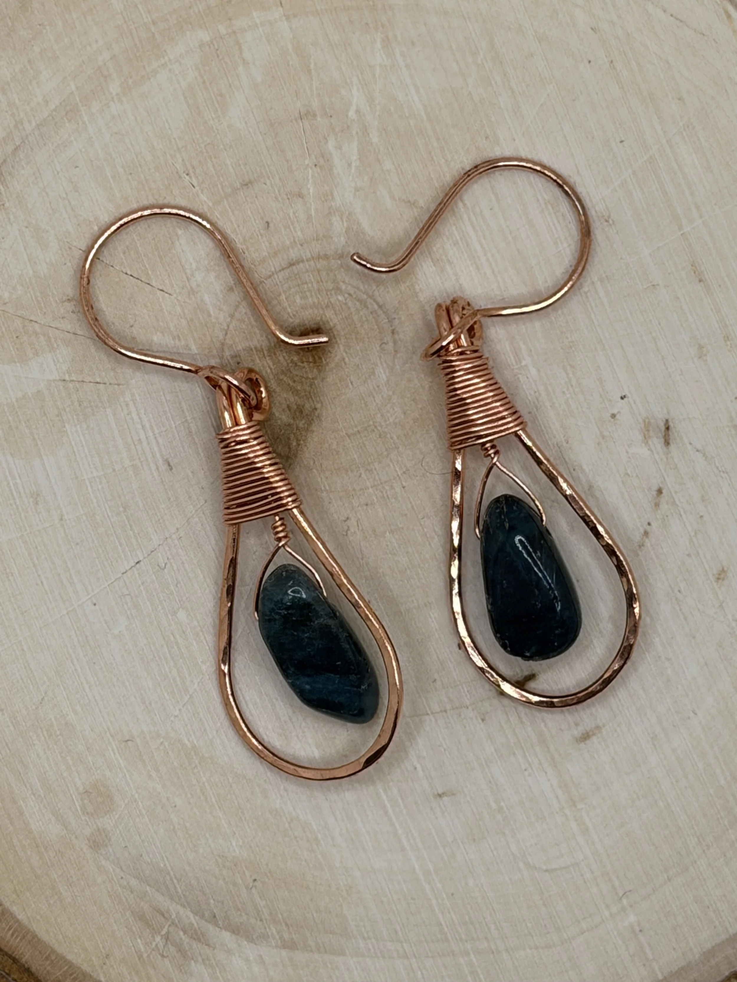Pair of handmade hammered copper teardrop earrings with coiled wire caps and deep blue tourmaline beads on a wooden slice