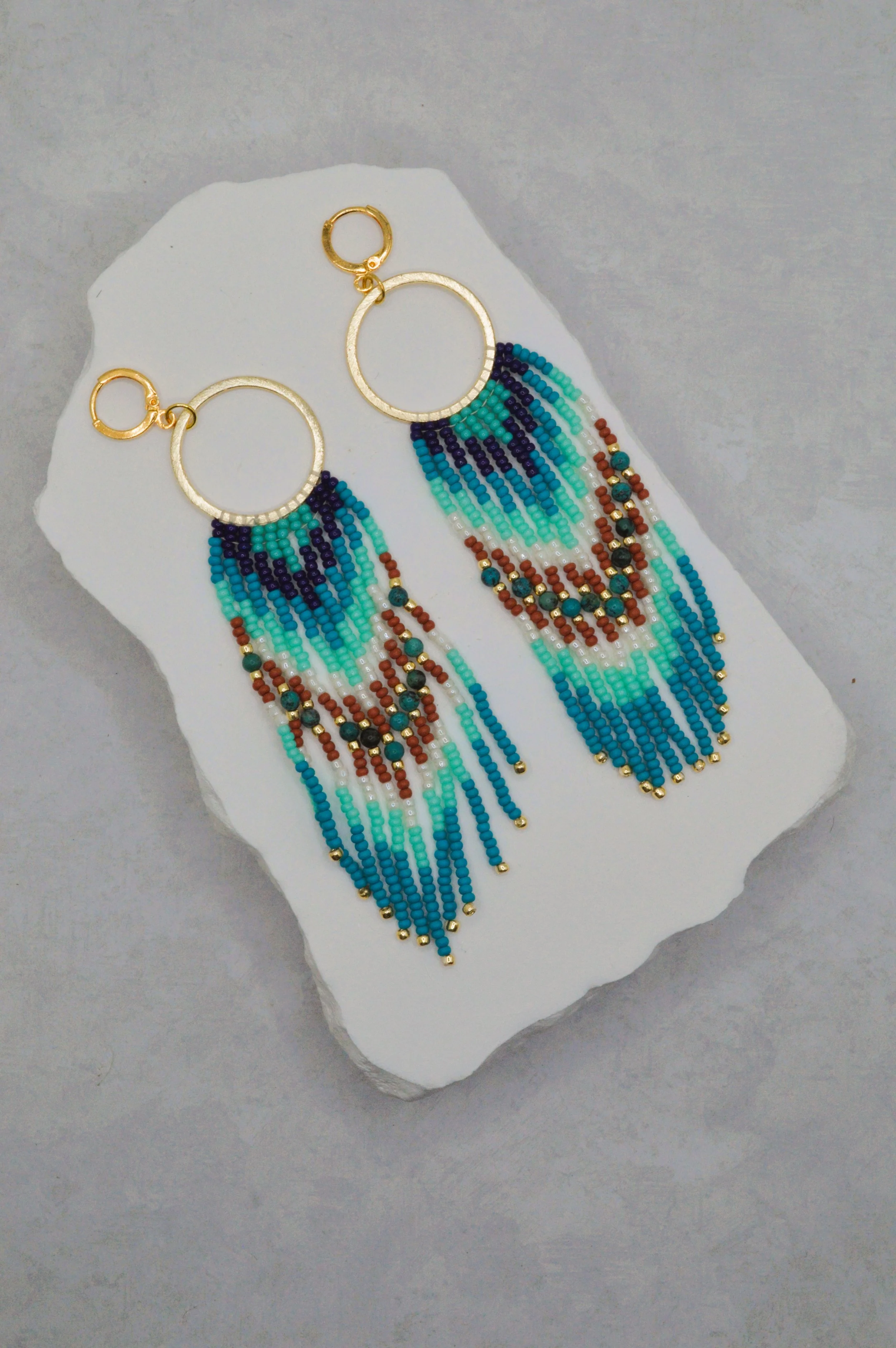 Pair of colorful beaded earrings with gold rings, hanging on a white piece of paper, placed on a gray surface.