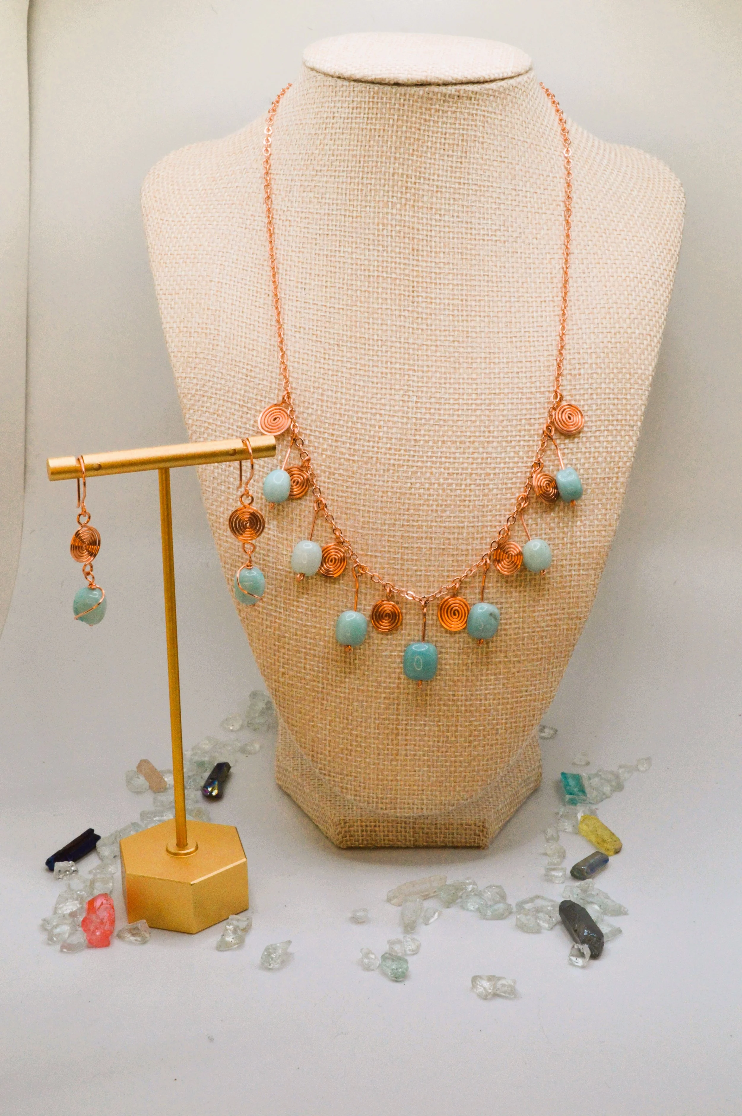 A jewelry display with a necklace and earrings featuring light blue beads and copper wire spirals, surrounded by loose crystals and stones, some on a gold stand.