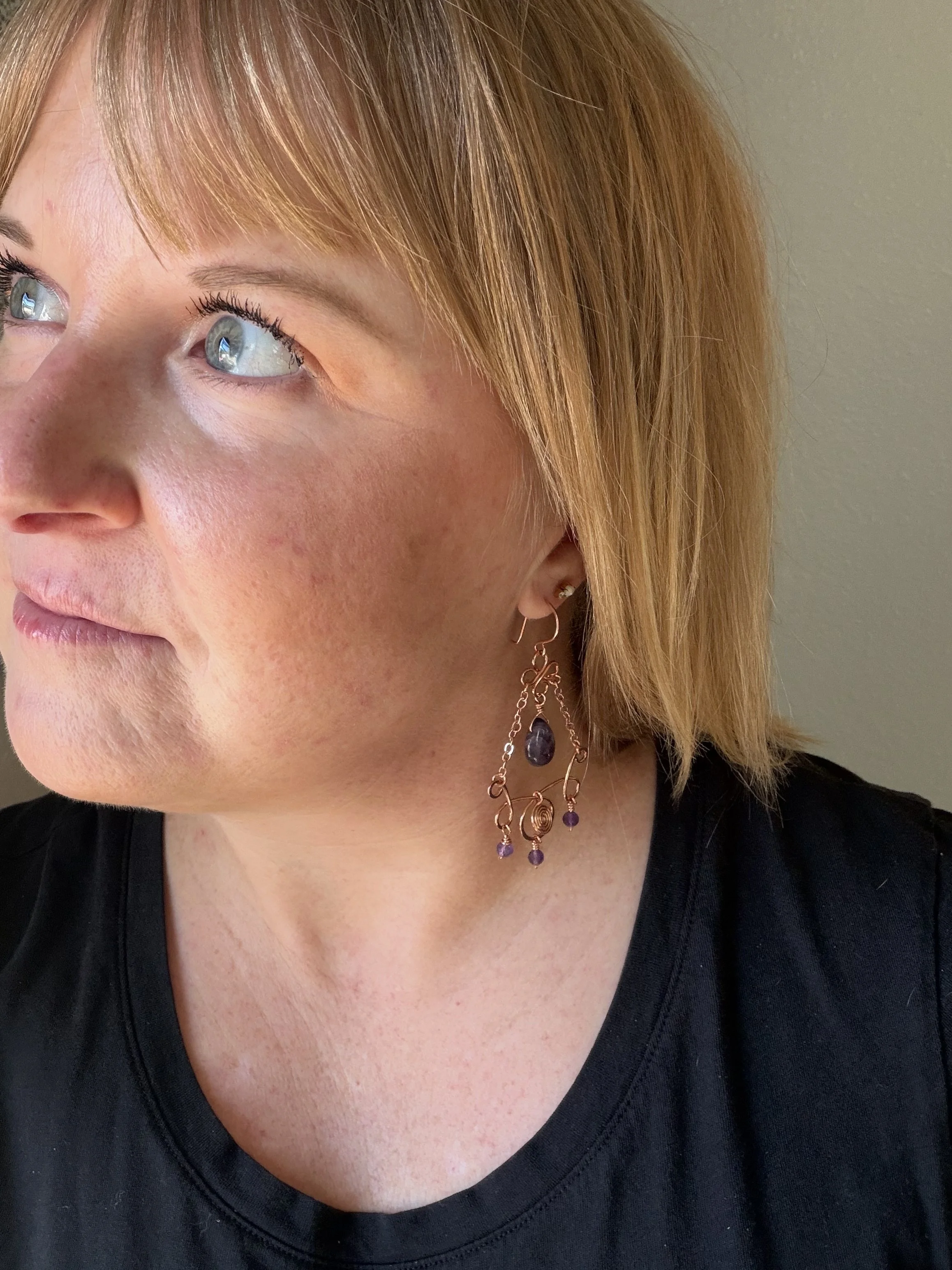 woman wearing Handmade copper chandelier earring worn on an ear, with scroll connectors, copper chains, a center spiral, an amethyst teardrop, and three small faceted amethyst beads.