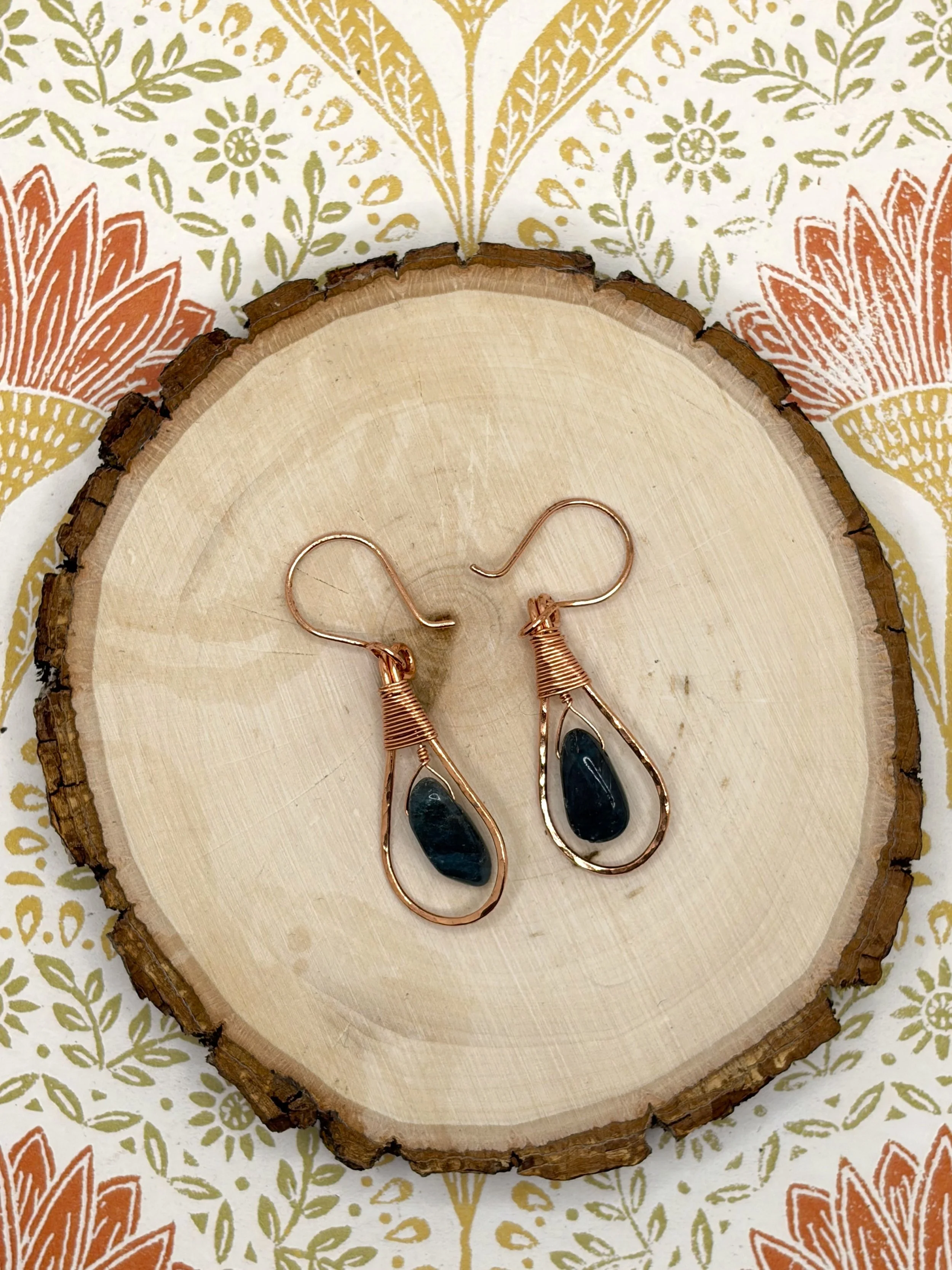 Pair of handmade hammered copper teardrop earrings with coiled wire caps and deep blue tourmaline beads on a wooden slice