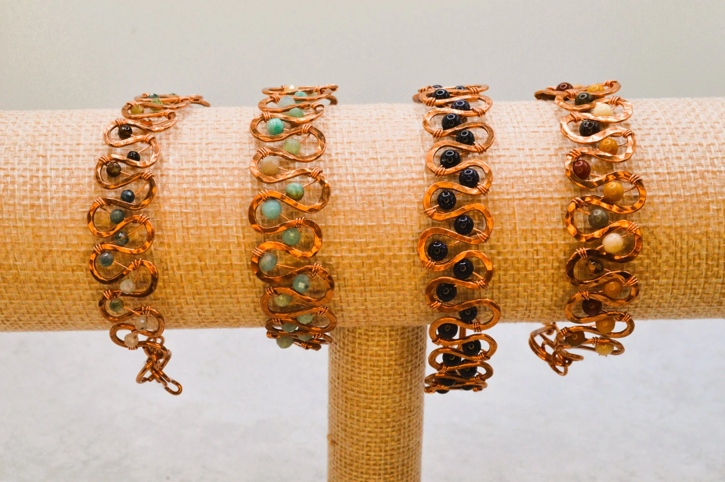 Multiple copper bracelets with various colored beads, displayed on a beige textured stand.