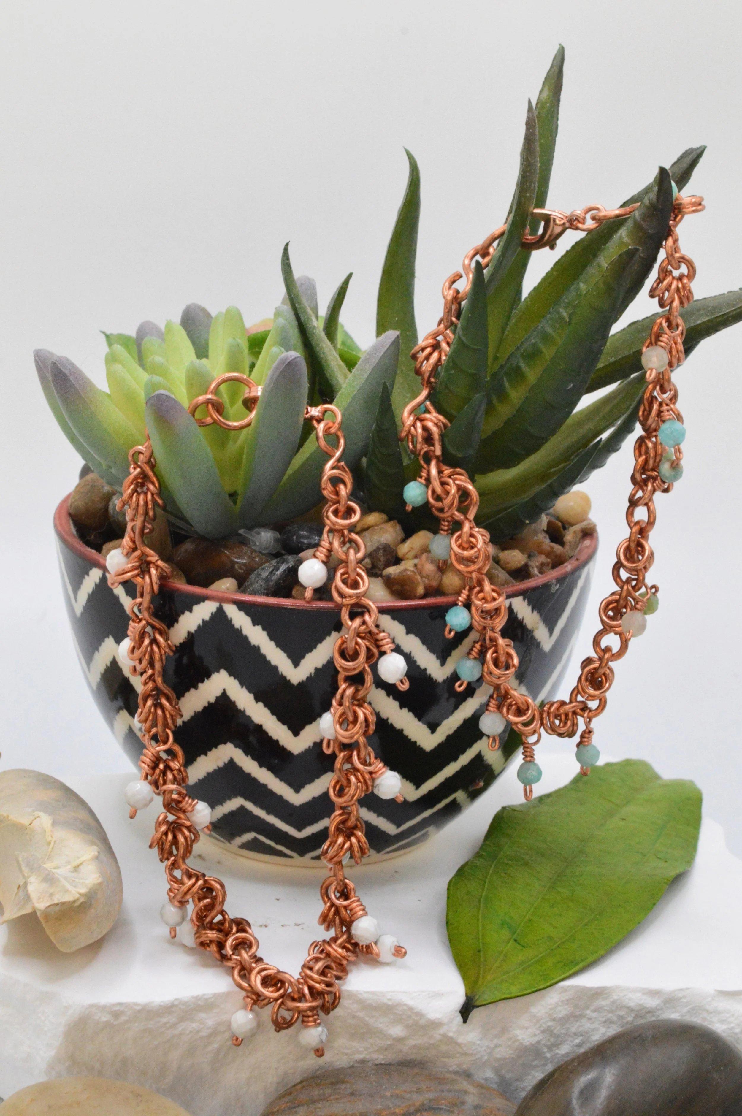 Close-up of a black and white patterned ceramic pot containing succulents with jewelry draped over the plants, surrounded by scattered stones and a green leaf.