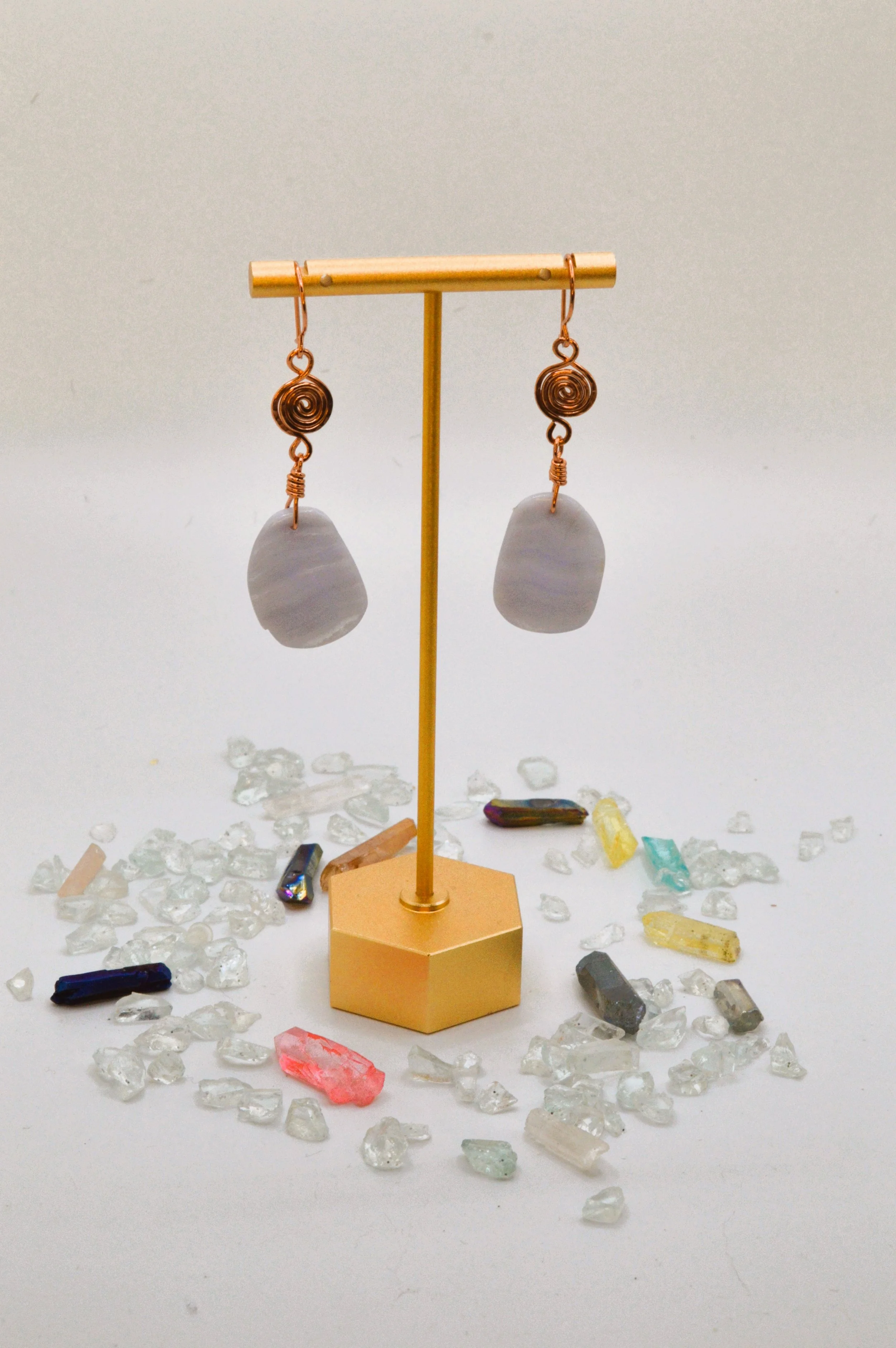 A pair of earrings with spiral copper wire designs and white stone pendants, displayed on a golden stand amidst scattered small clear and colored crystals on a white background.