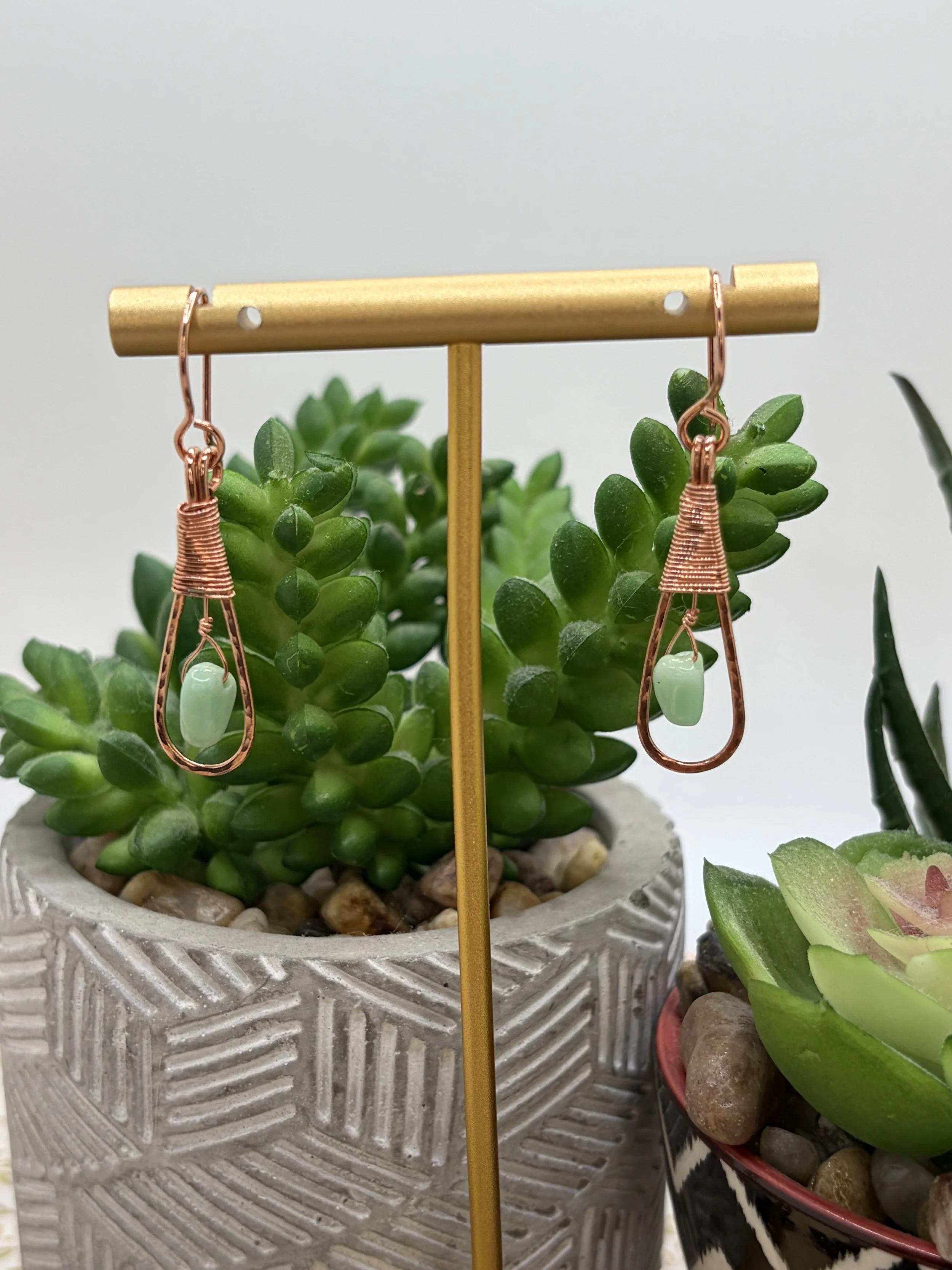 Pair of handmade hammered copper teardrop earrings with coiled wire caps and mint-green chrysoprase beads hanging from a gold display stand in front of succulents.
