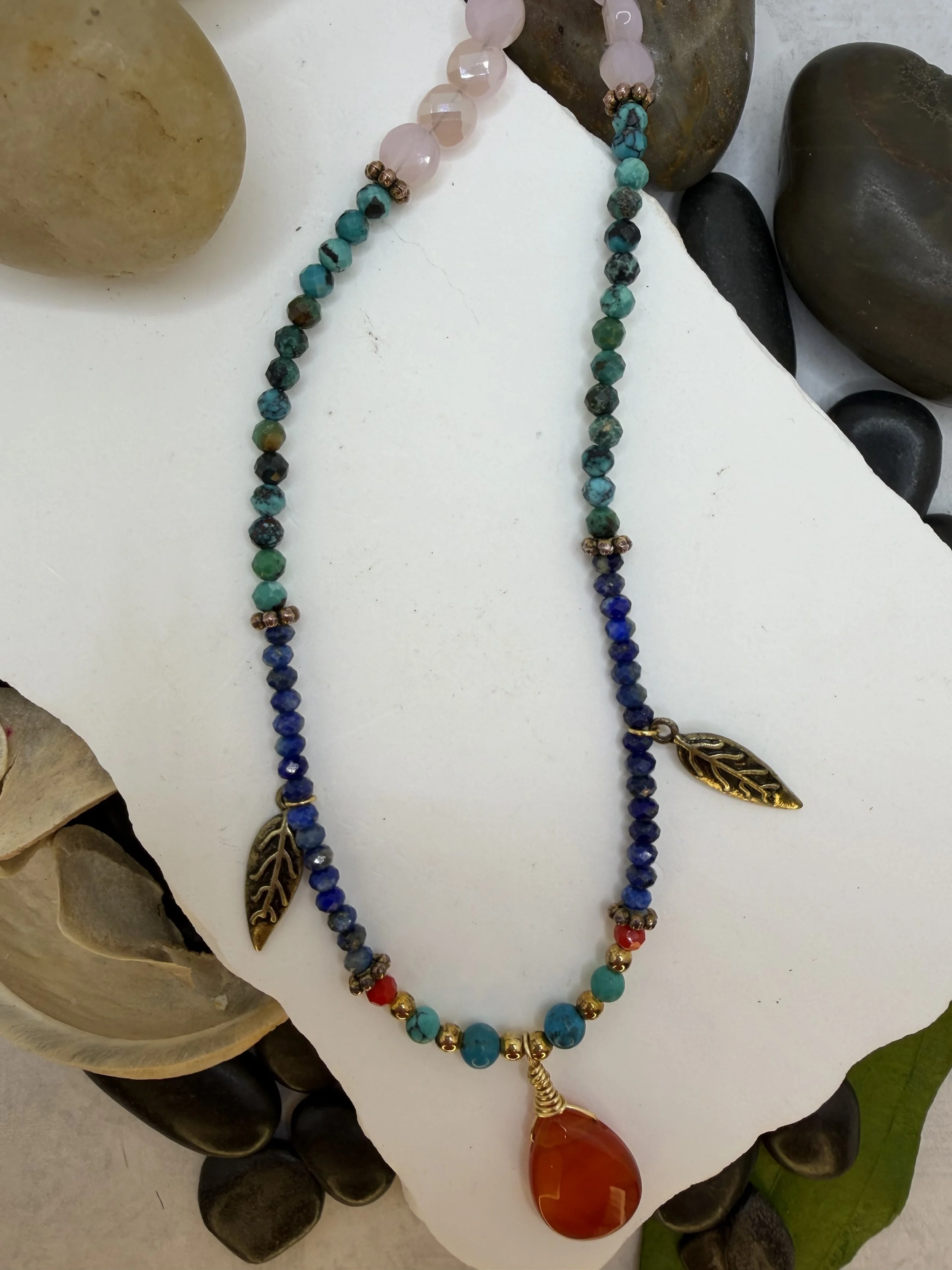 Beaded necklace with carnelian pendant, turquoise, lapis lazuli, pink glass beads, and brass feather charms on a lobster claw clasp.