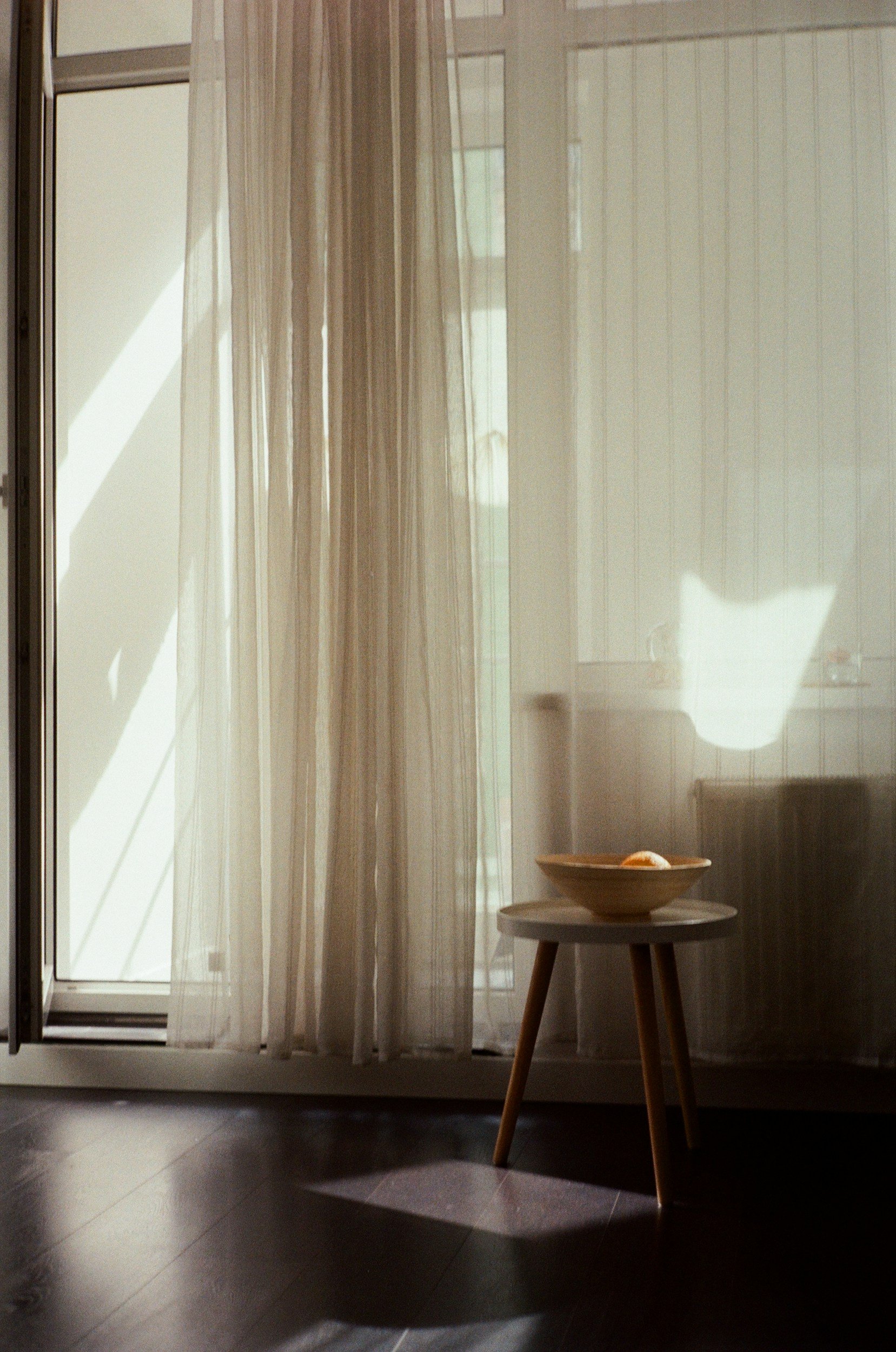 A wooden stool with three legs and a round top, placed in front of a sheer curtain and glass door. The sunlight filters through the curtain, creating shadows on the floor.
