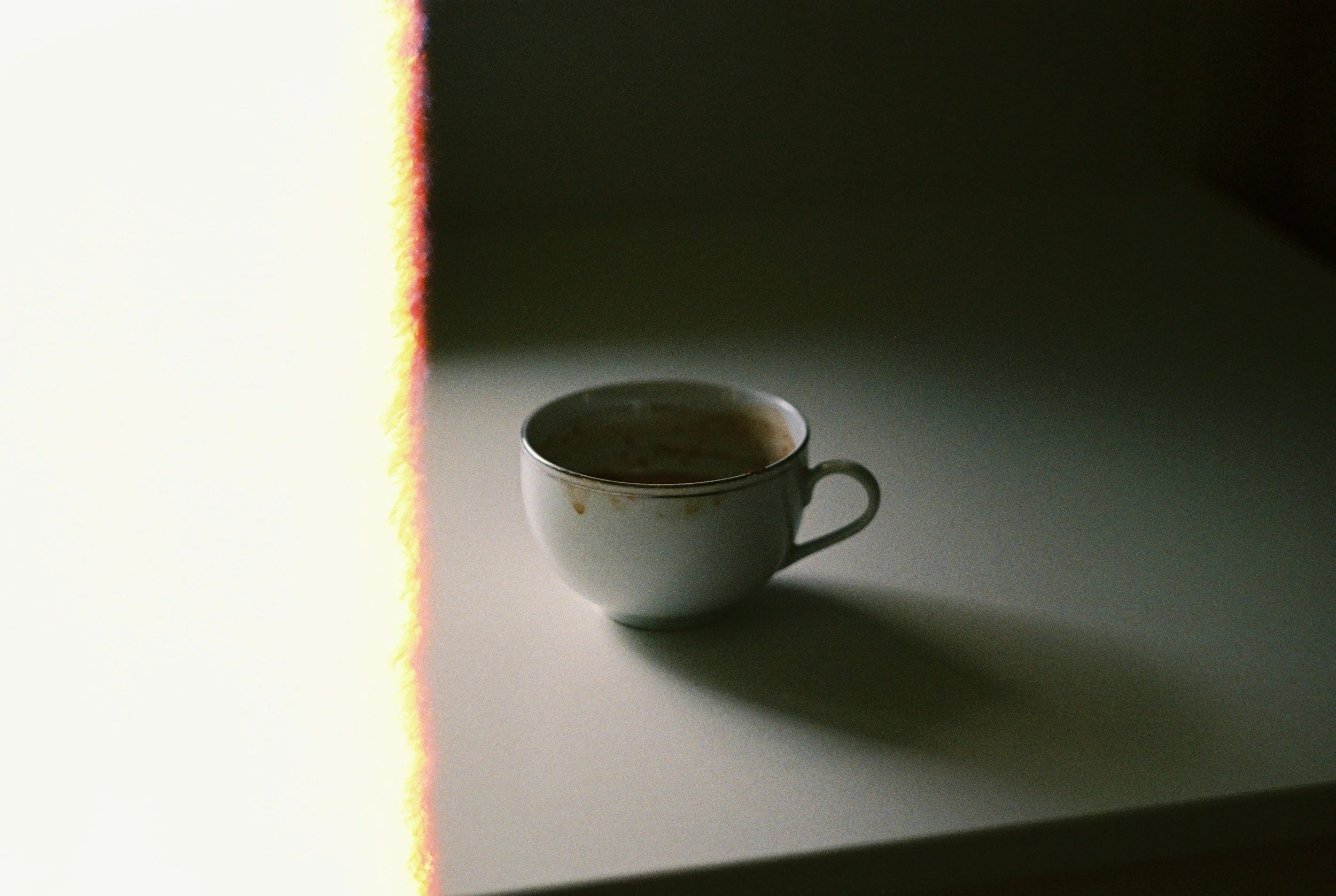 A white coffee cup with a floral pattern on a white surface, with a dark background to the right and a bright, colorful light streak on the left side of the image.