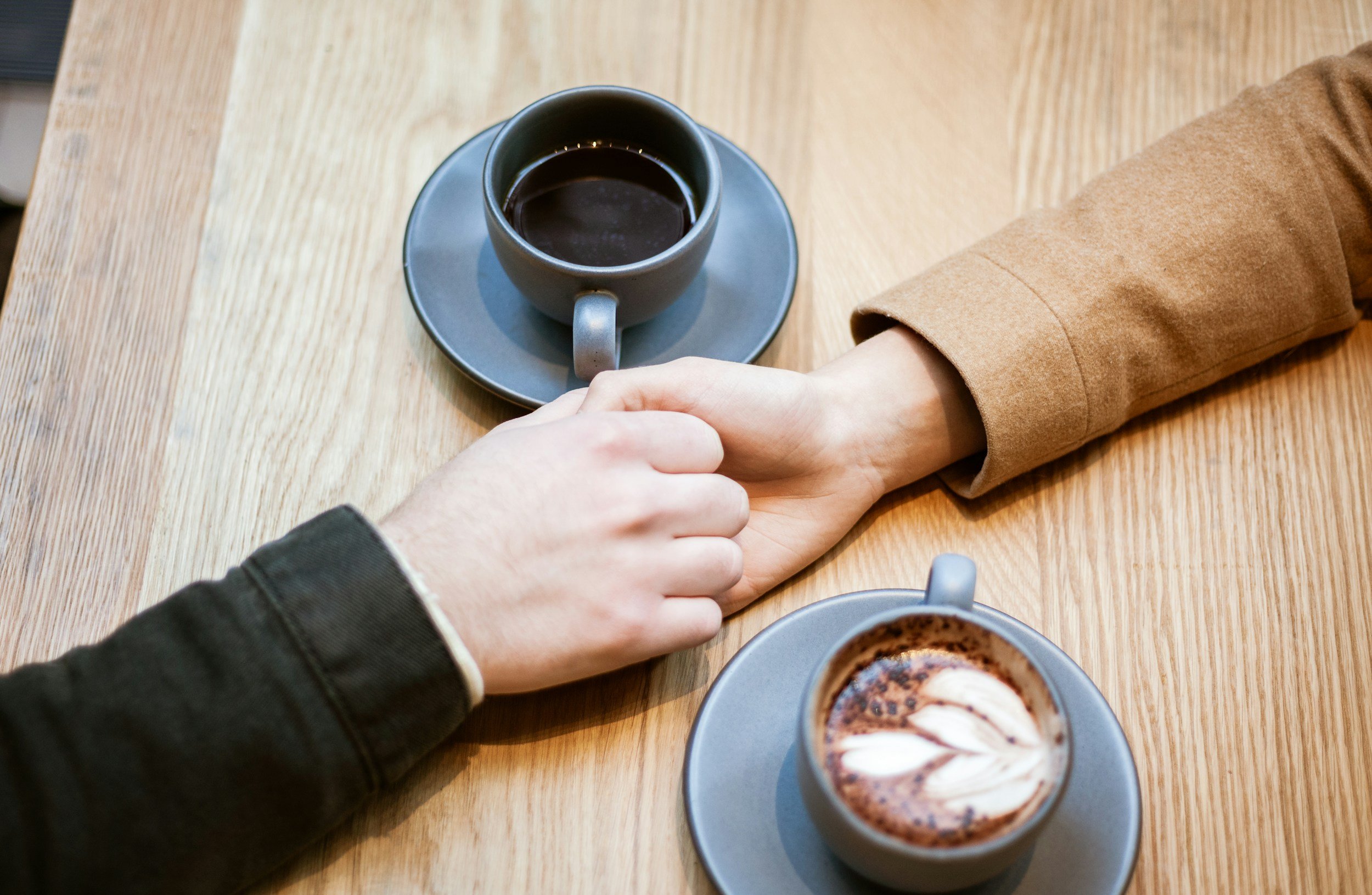 A person wearing a black hoodie is holding hands with another person wearing a tan coat over a light-colored shirt across a wooden table. On the table, there are two gray cups, one filled with black coffee and the other with a hot chocolate topped with whipped cream and chocolate shavings.