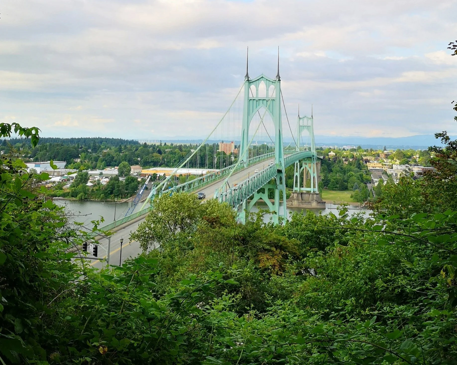 A view of the St. Johns Bridge in Portland, Oregon, with its distinctive Gothic-style towers and suspension cables, surrounded by lush green trees and a river in the foreground, under a partly cloudy sky.