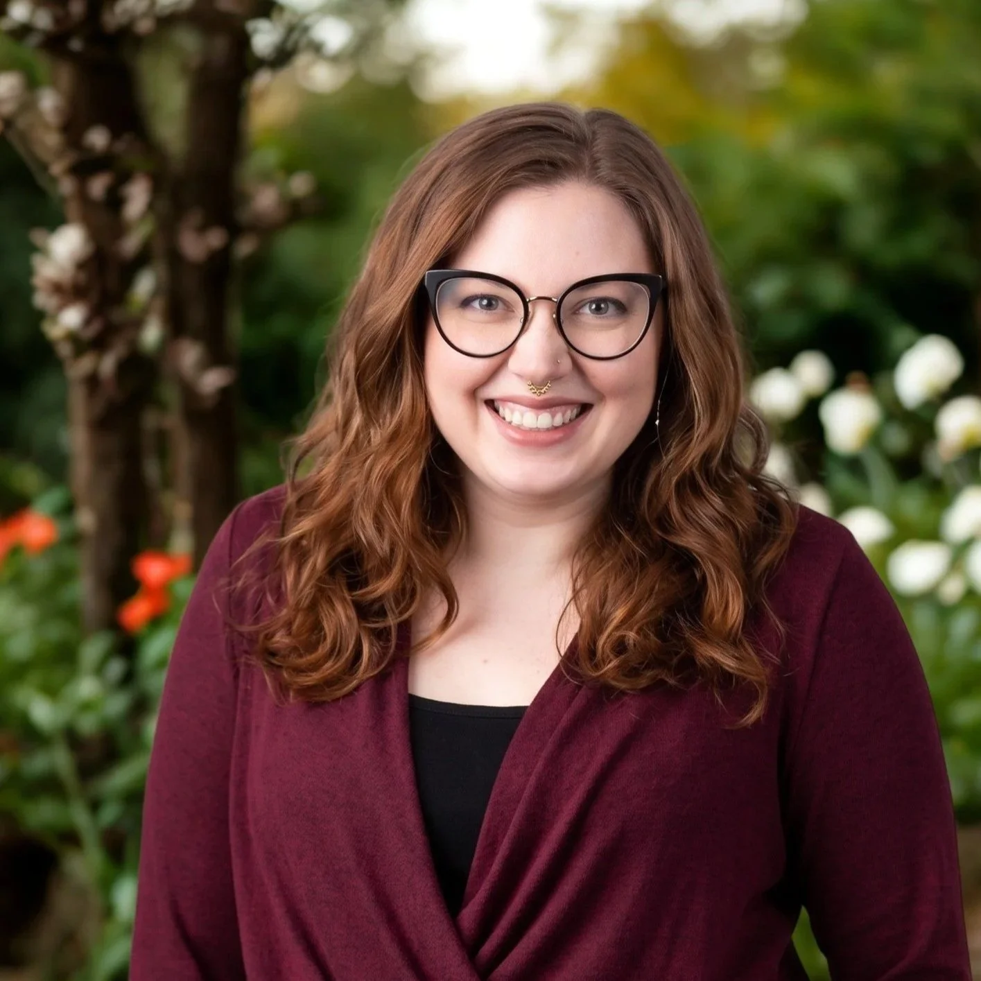 A woman with long, wavy, auburn hair, wearing glasses and a nose ring, smiling, outdoors with greenery and flowers in the background.