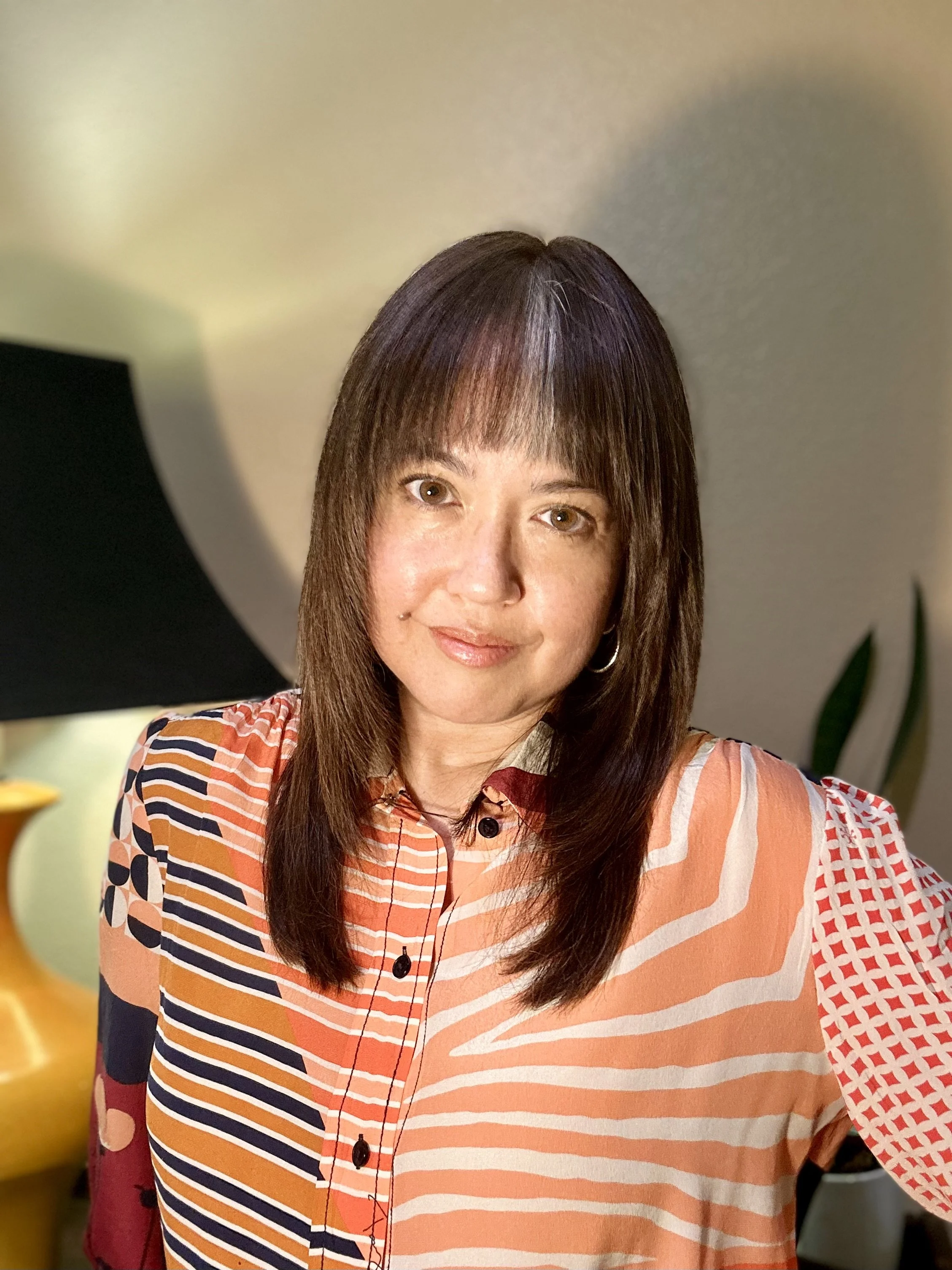 A woman with brown hair and brown eyes smiling at the camera. She is wearing a colorful, striped shirt with various patterns. The background includes a green plant and a blurred yellow object to the left.