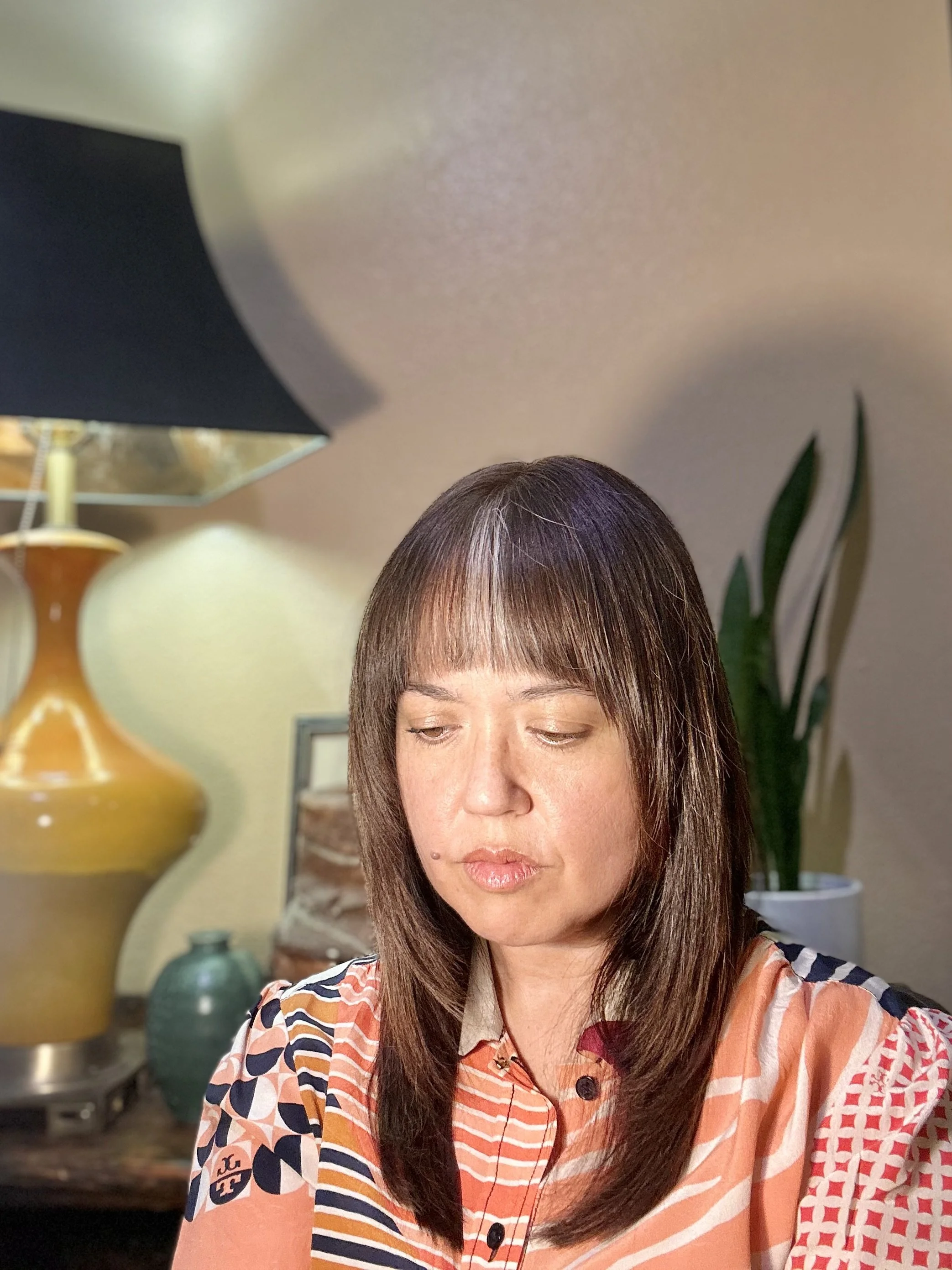 A woman with brown hair and bangs, wearing a colorful patterned top, sitting indoors with a background of decorative vases and a plant.