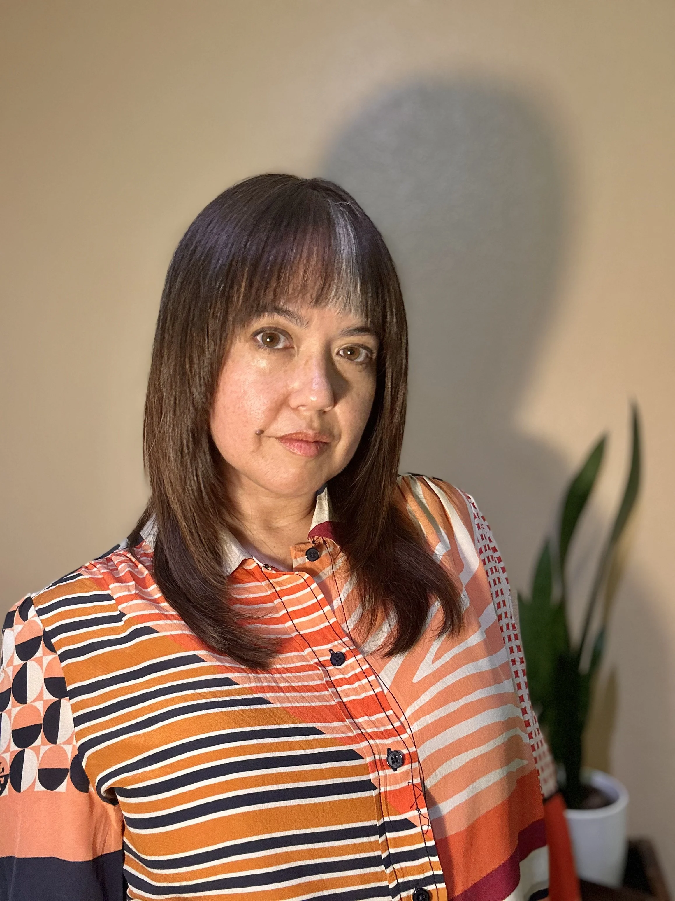 A woman with shoulder-length brown hair and hazel eyes, wearing a multicolored striped blouse with a collar and buttons, stands in front of a beige wall with a green plant in a white pot visible in the background.