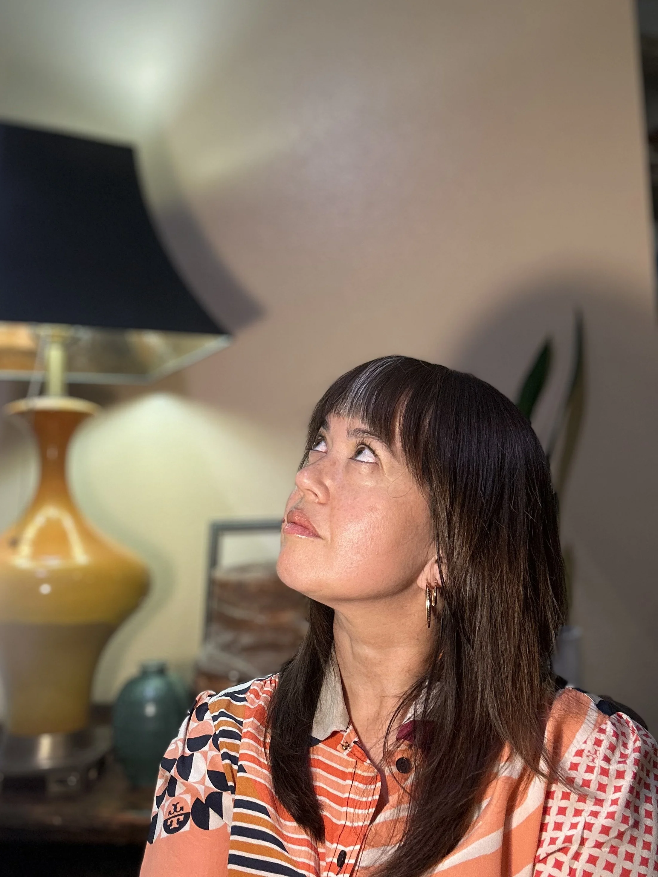A woman with dark brown hair and hoop earrings looks upward in a room with a tan wall, large yellow and white vases, and a lamp in the background.
