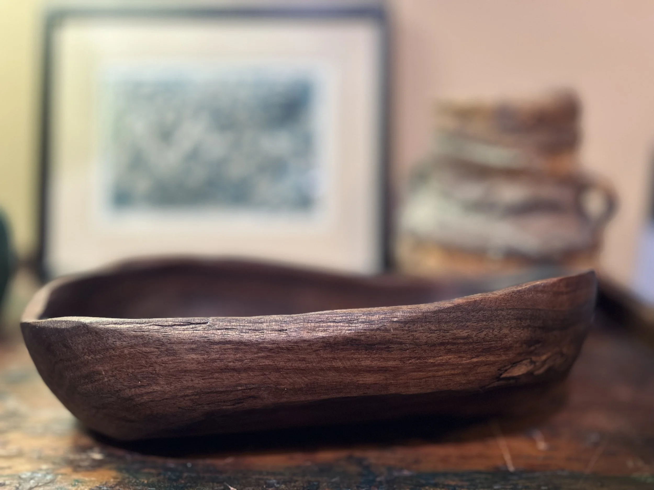 Close-up of a small wooden bowl on a wooden surface, with an out-of-focus background including a picture frame and shelves.