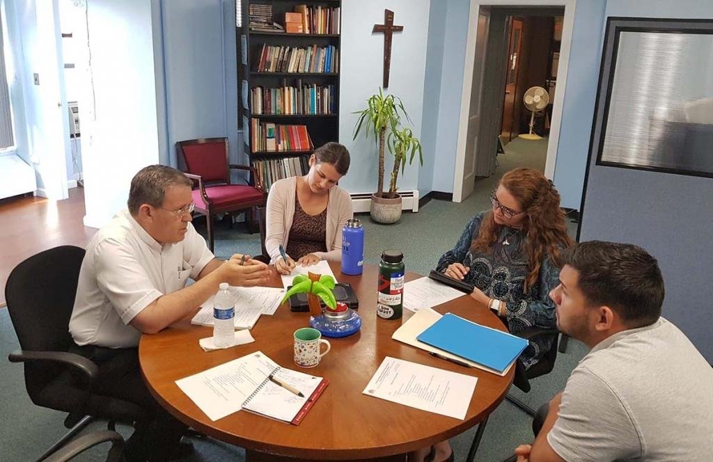 Four people sitting around a wooden table in a room with bookshelves, a crucifix, a potted plant, and a closet, engaged in a discussion or meeting with papers, notebooks, and drinks on the table.