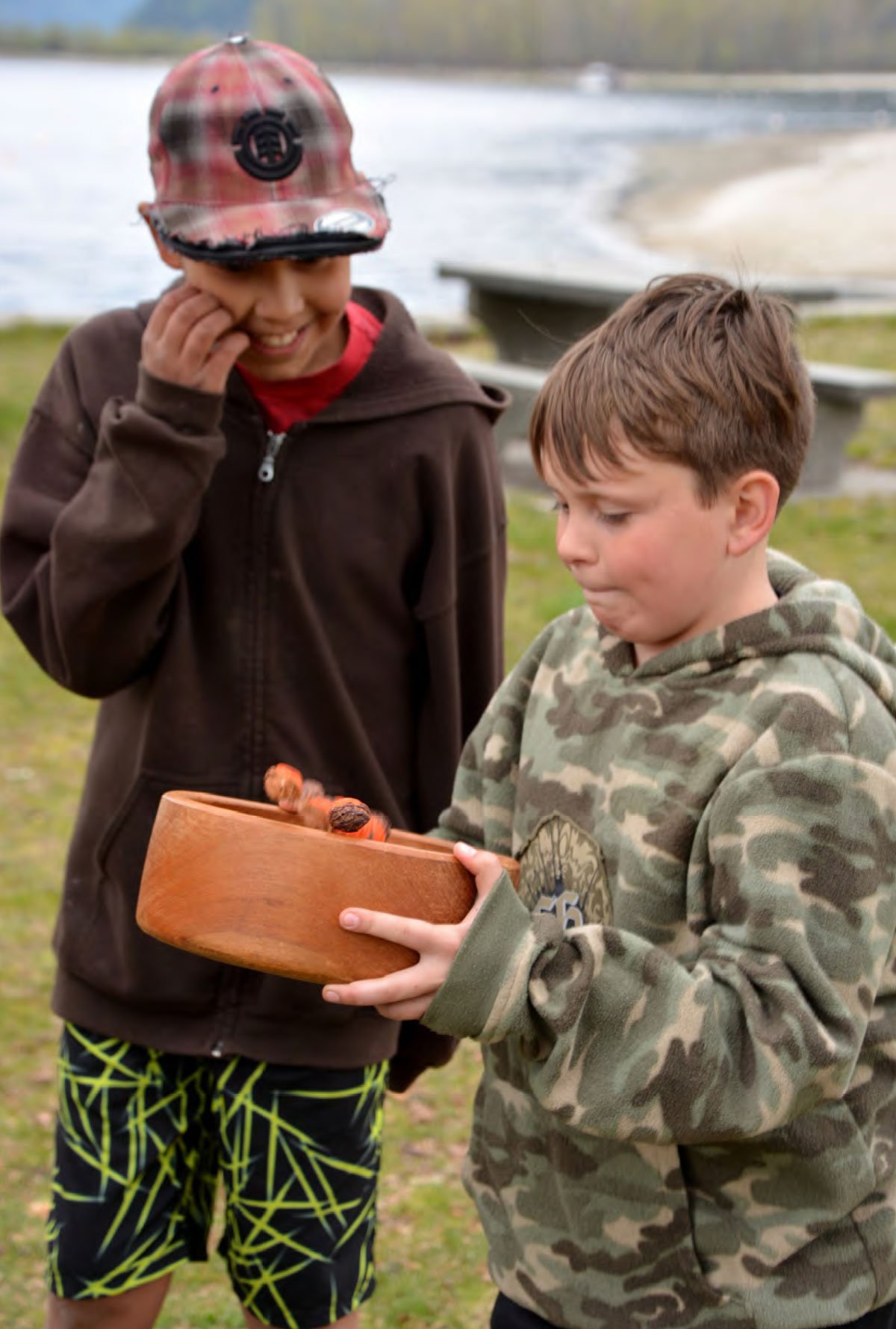 Two boys outdoors near a body of water, one wearing a plaid cap and a brown hoodie, smiling and touching his face, and the other in a camouflage hoodie holding a round wooden container with insects inside, appearing curious.