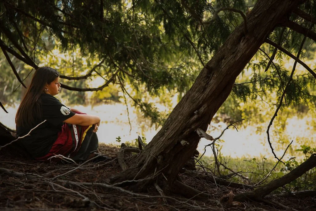 A girl sitting under a large tree in a forested area, gazing at the water nearby.