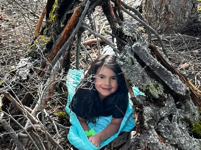 A young girl lying inside a makeshift tent made of branches and wood in a forest, smiling at the camera.