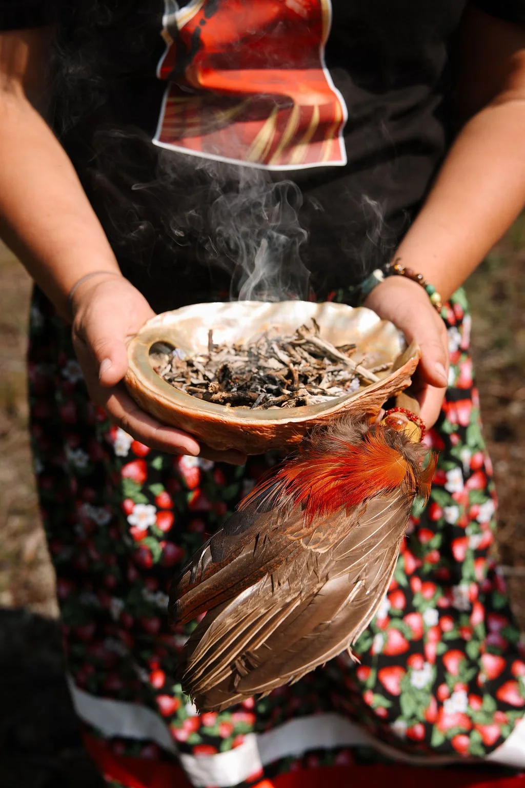 Person holding a smoking shell filled with ashes, adorned with a colorful feathered accessory, outdoors with floral patterned skirt visible.