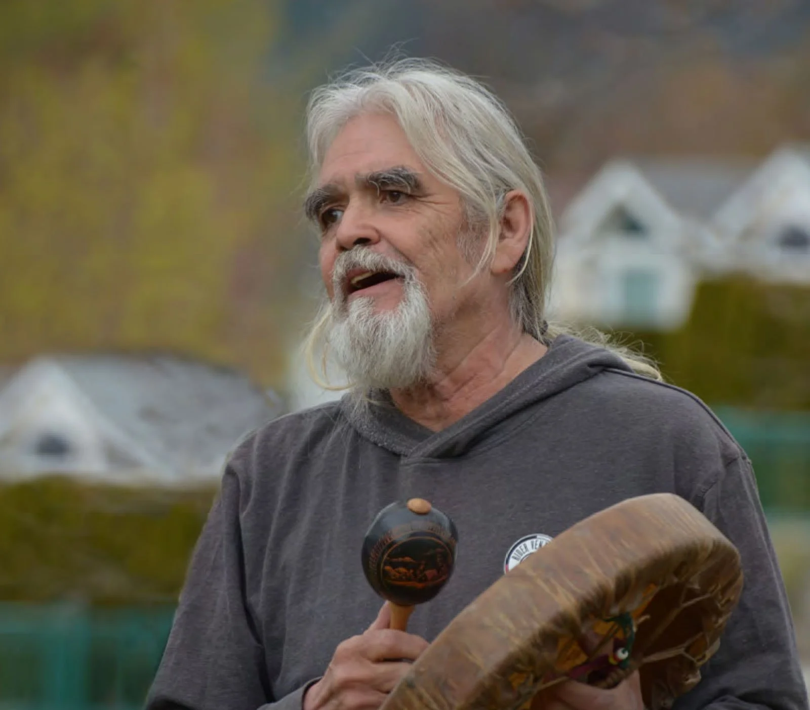 Indigenous Elder with long gray hair and beard, wearing a gray hoodie, holding a drum in one hand and a mallet in the other, outdoors with blurred houses and trees in the background.