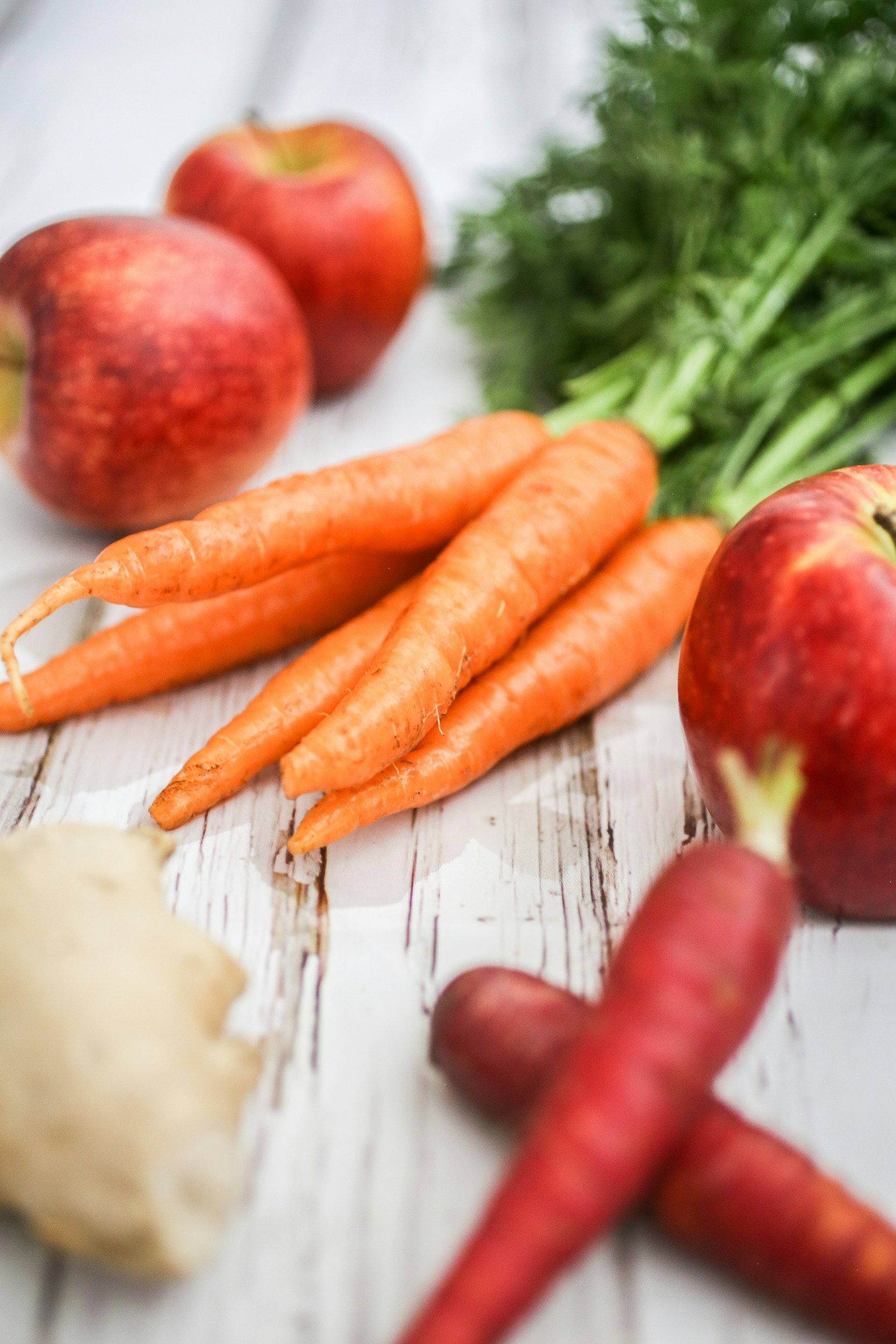 Fresh carrots, apples, and a ginger root on a white wooden surface.