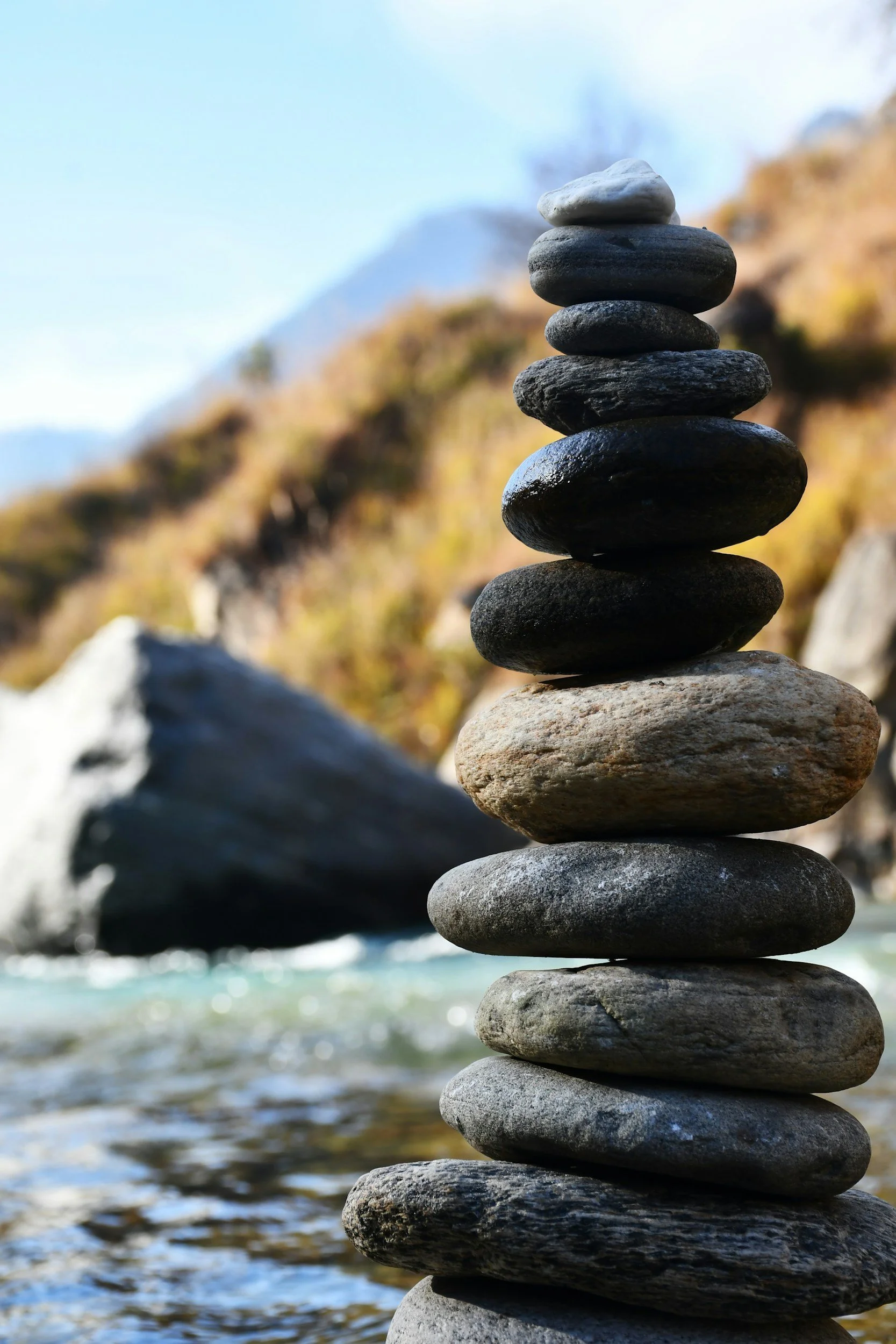A balanced tower of smooth stones stacked on top of each other near a body of water with a rocky background and a mountain in the distance.