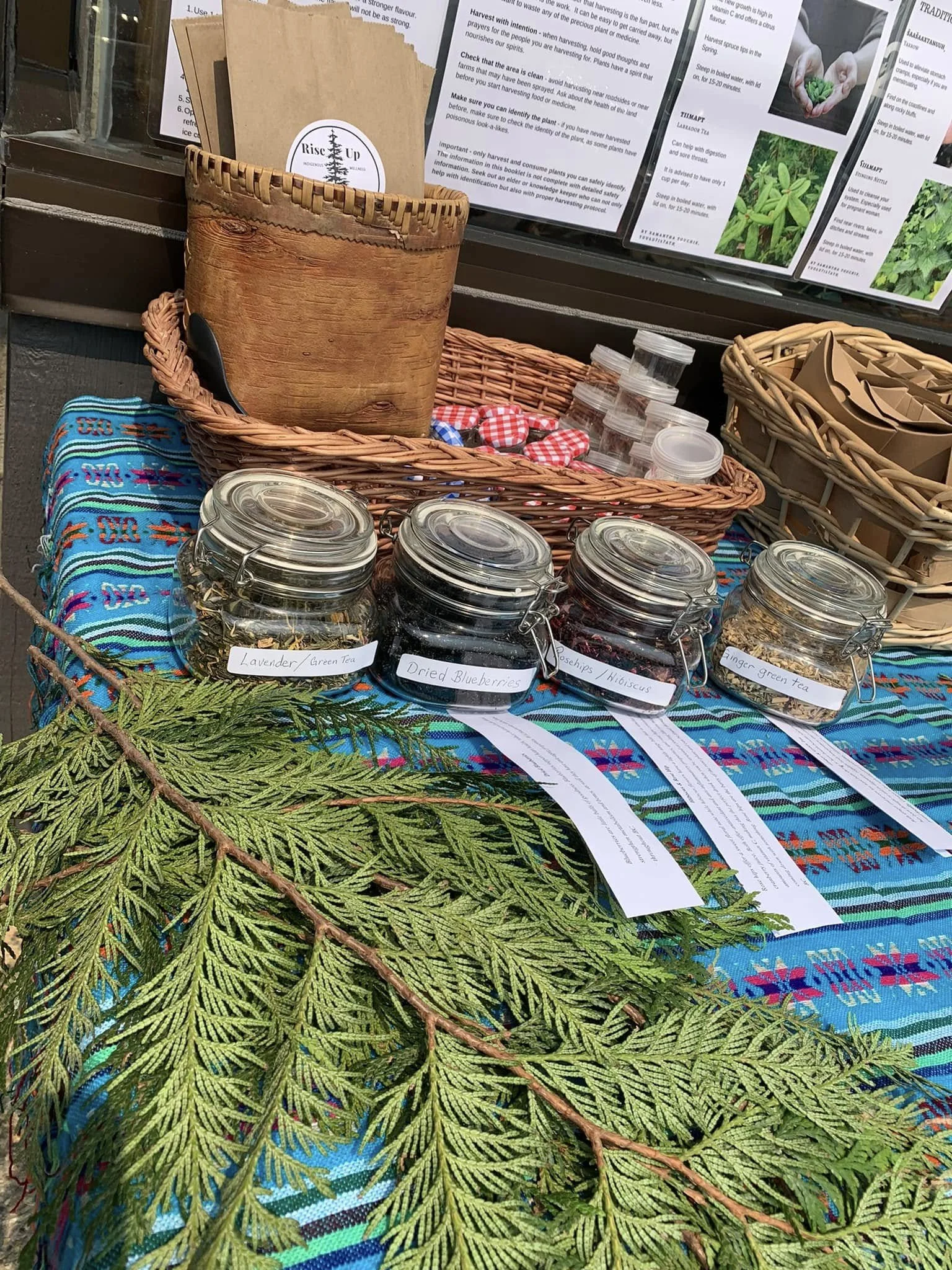 A display table with jars of dried herbs and tea labeled lavender, blueberry, and green tea, decorated with green fern leaves, patterned cloth, and informational pamphlets behind.