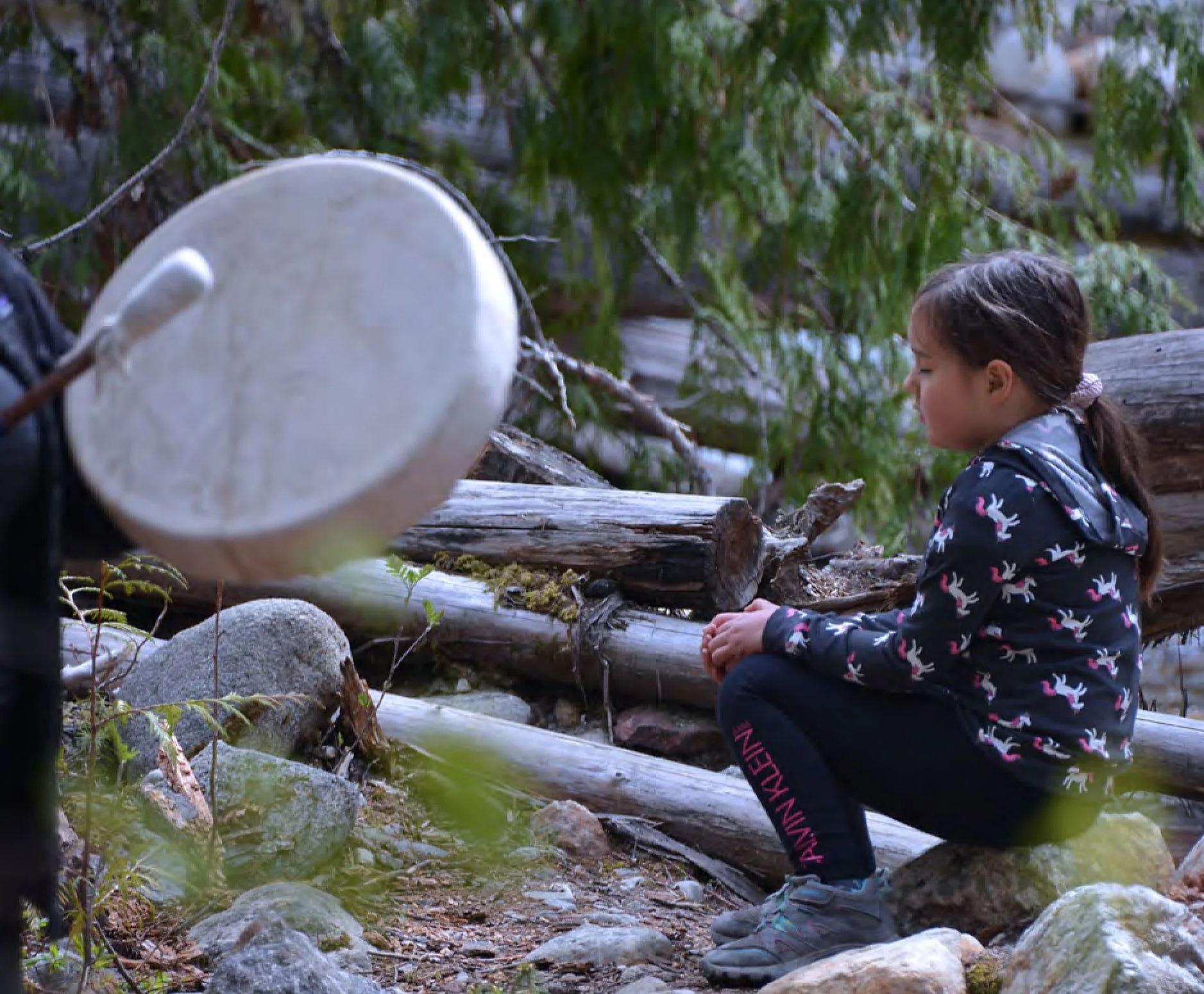 A young girl with brown hair tied back, sitting outdoors on a large log near a stream, surrounded by rocks and green foliage, looking down. A hand drumming a drum in the foreground on the left.