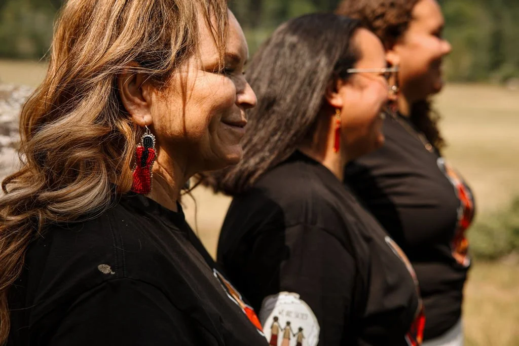 Three women standing outdoors, side profile, smiling, wearing black shirts and red earrings, with trees and grass in the background.