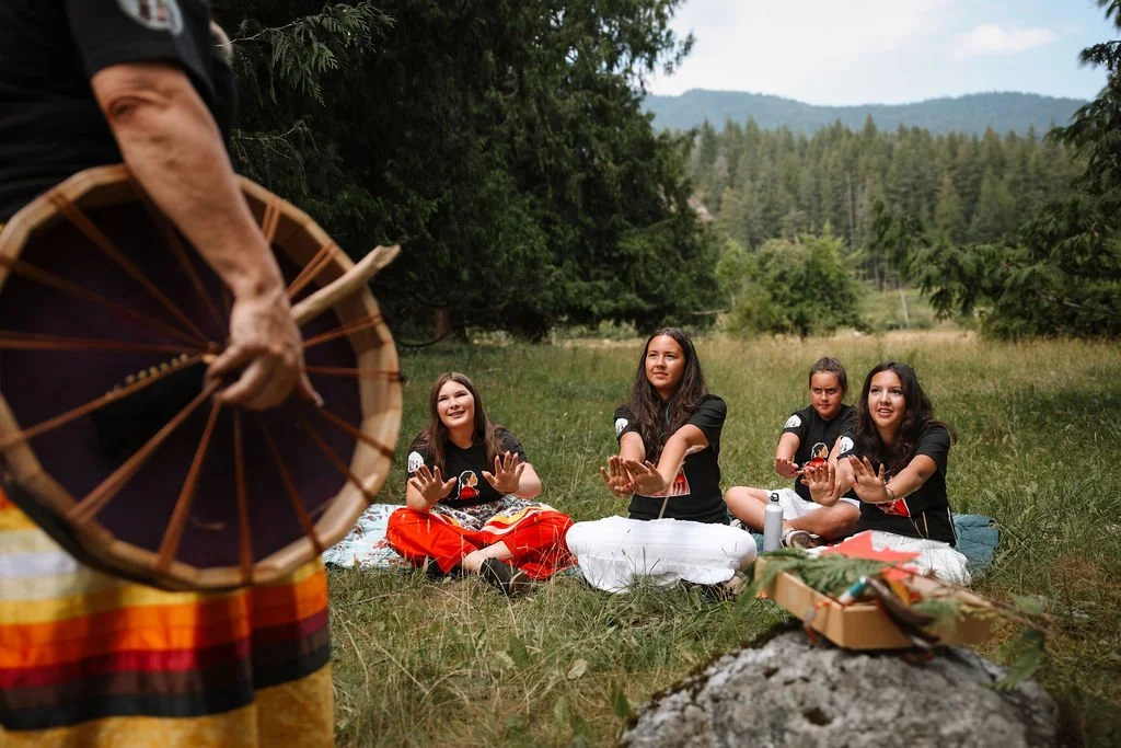Four young women sitting cross-legged on the grass, processions and applying traditional hand gestures as an instructor plays a drum in the foreground during an outdoor cultural or spiritual event.