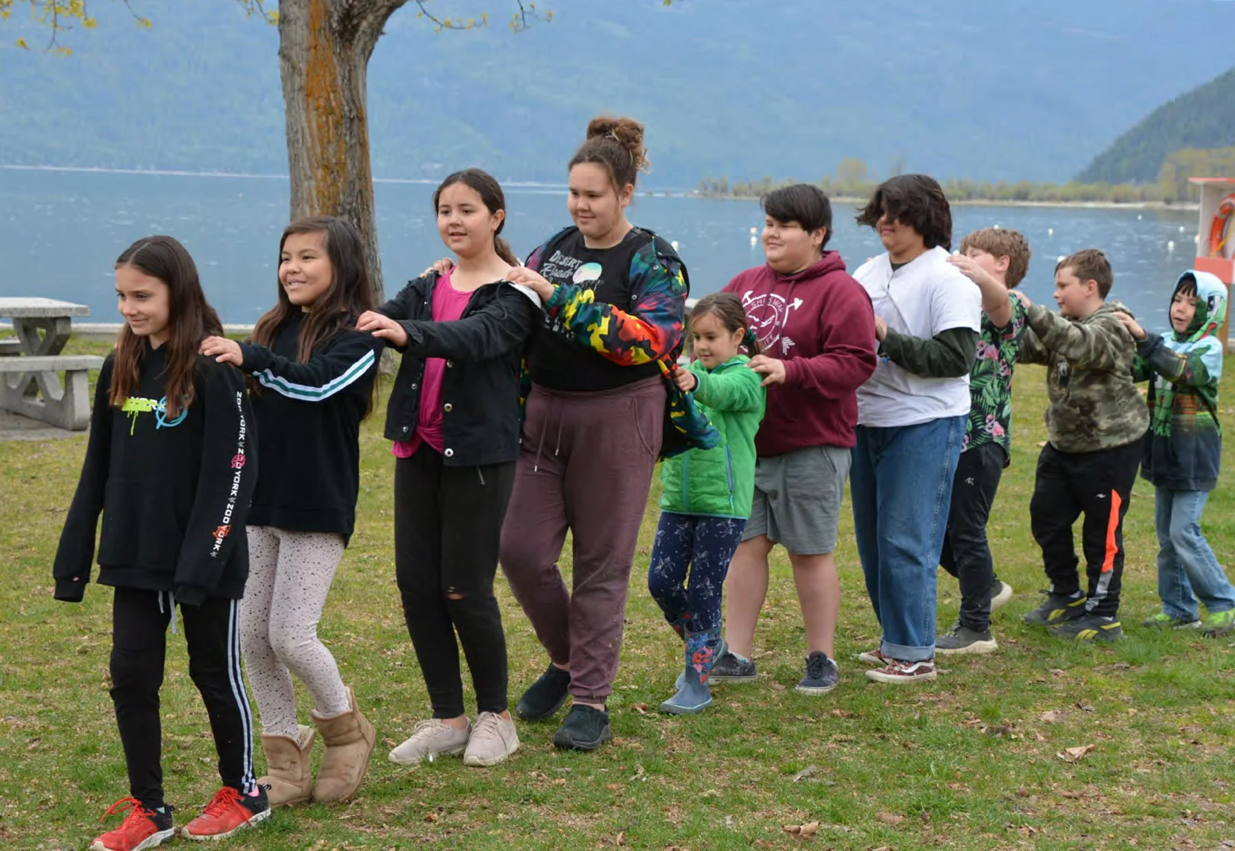 A group of children and a young woman are standing in a line outdoors, participating in a line dance near a lake with mountains in the background.