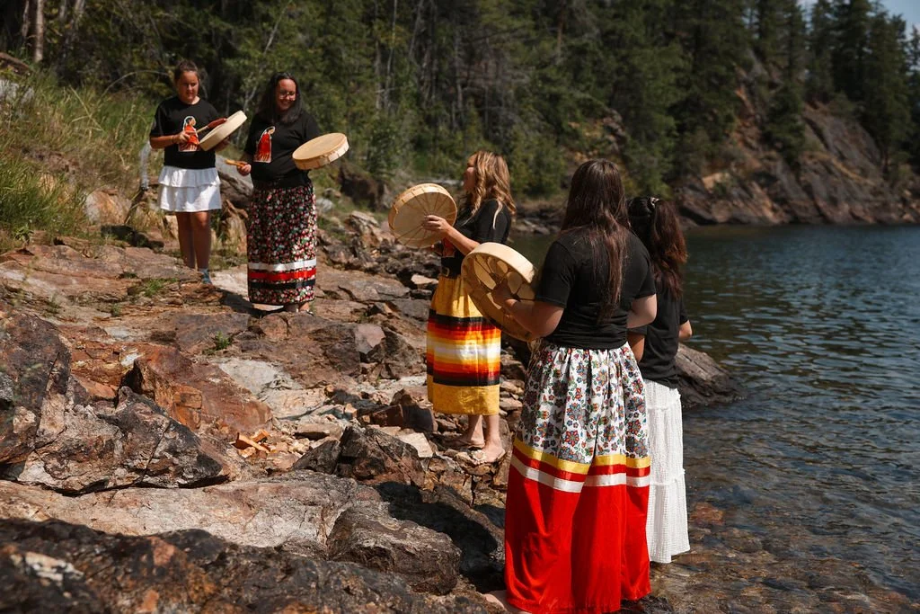 Group of women on a rocky lakeshore, holding and playing drums, dressed in traditional clothing, with trees and water in the background.