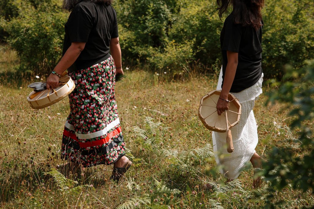 Two women walking in a grassy field with trees in the background, holding woven baskets.