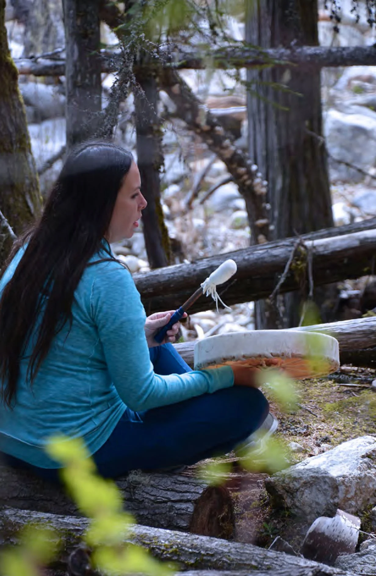 A woman sitting outdoors on a log, holding a a drum in front of her, surrounded by trees and rocks.