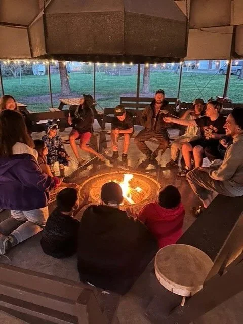 A group of people, including children and adults, sitting around a campfire inside a screened pavilion, with some people roasting marshmallows and enjoying conversations.