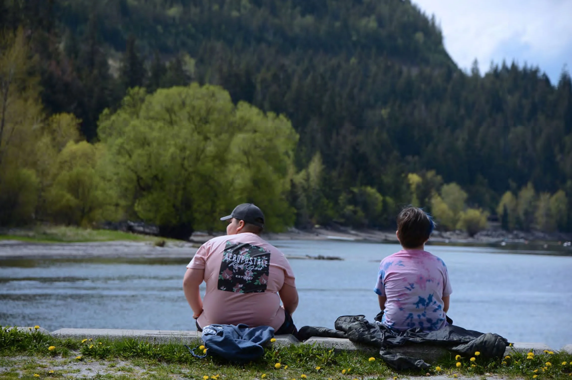 Two children are sitting on rocks by a river, facing the water with mountains and trees in the background, during daytime.