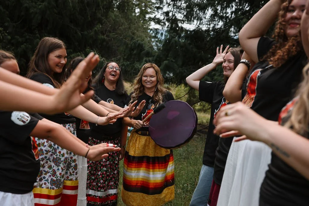 Group of women outdoors in a circle, smiling and clapping, some women holding a colorful drum or shamanic instrument, surrounded by trees.