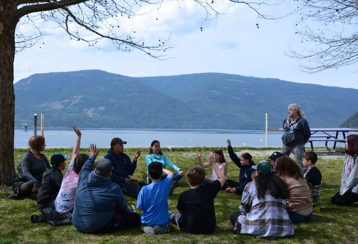 A group of people by a rocky shoreline with a large body of water in the background, mountains in the distance under a blue sky with wispy clouds.