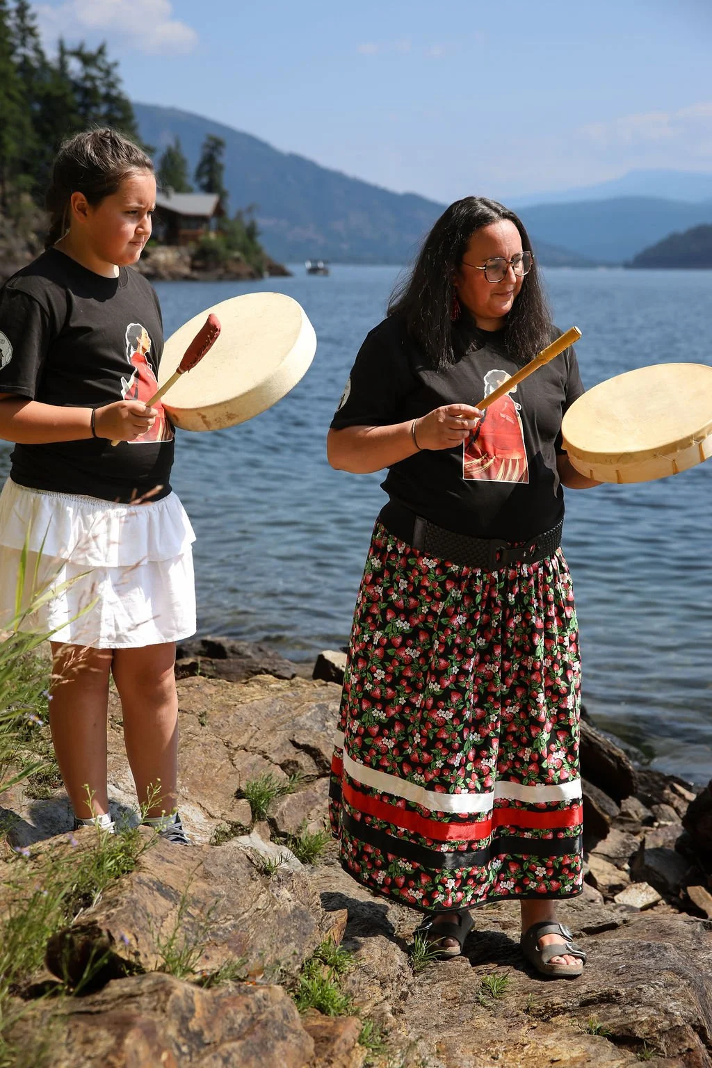 Two women stand by a lake, holding large wooden drums and drumsticks, near rocks with a backdrop of mountains and a partly cloudy sky.