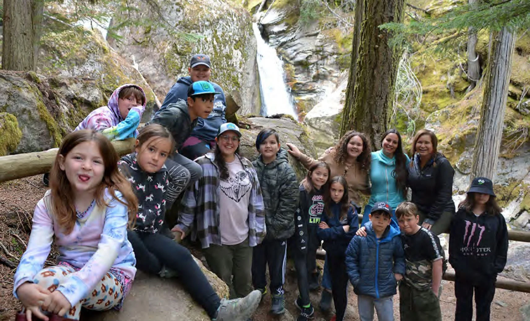 A group of children and adults hiking in a forest with a waterfall in the background.