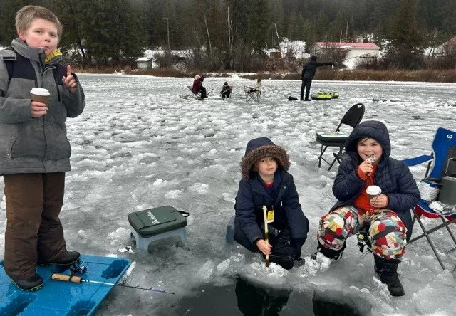 Children ice fishing on a frozen lake, with fishing gear and chairs, snow-covered trees in the background.