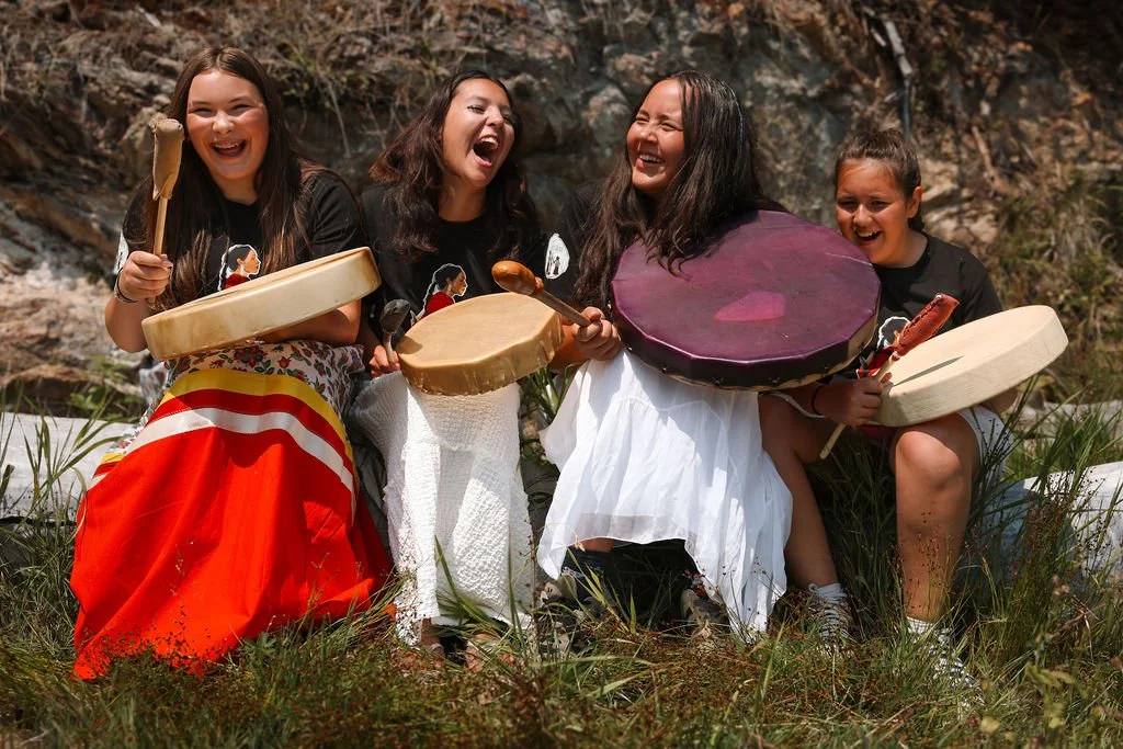 Four girls laughing and playing drums outdoors next to a river with rocks and grass.